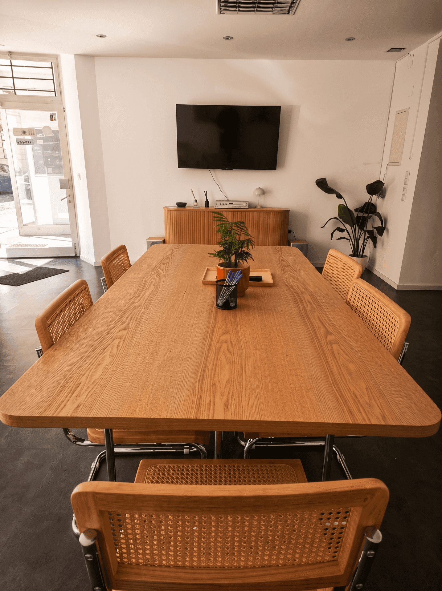 Großer Holztisch mit Rattan-Stühlen in einem hellen Konferenzraum mit Wandfernseher.