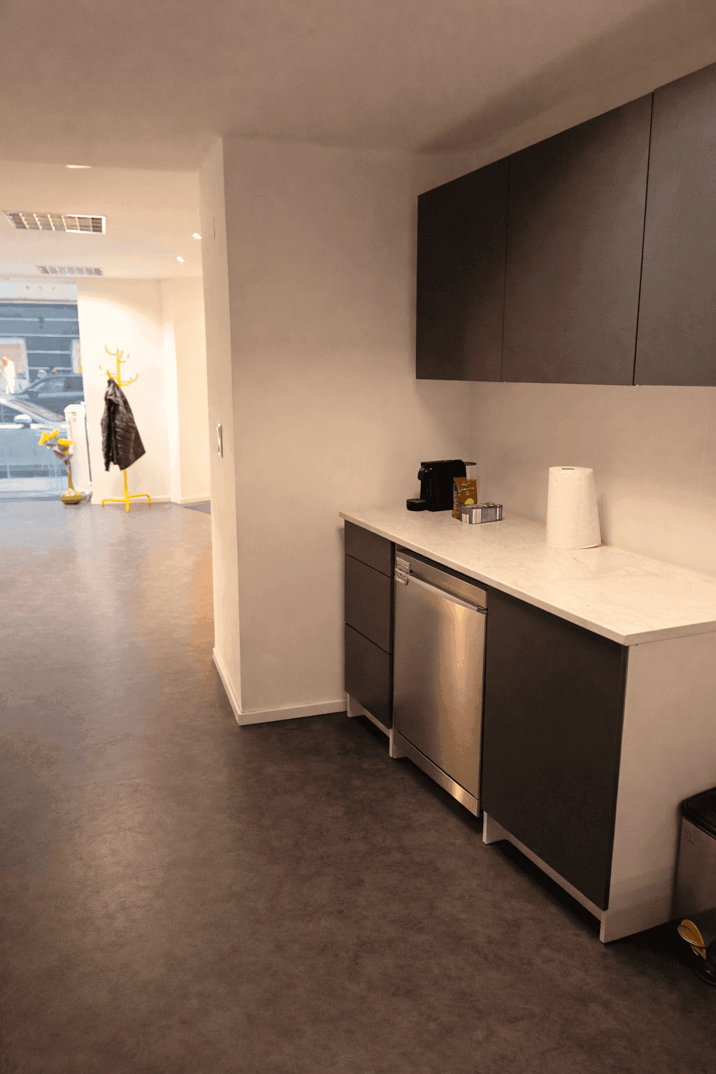 Kleine Küchenzeile mit Spülmaschine, Kaffeemaschine und dunklen Schränken in einem Büro.