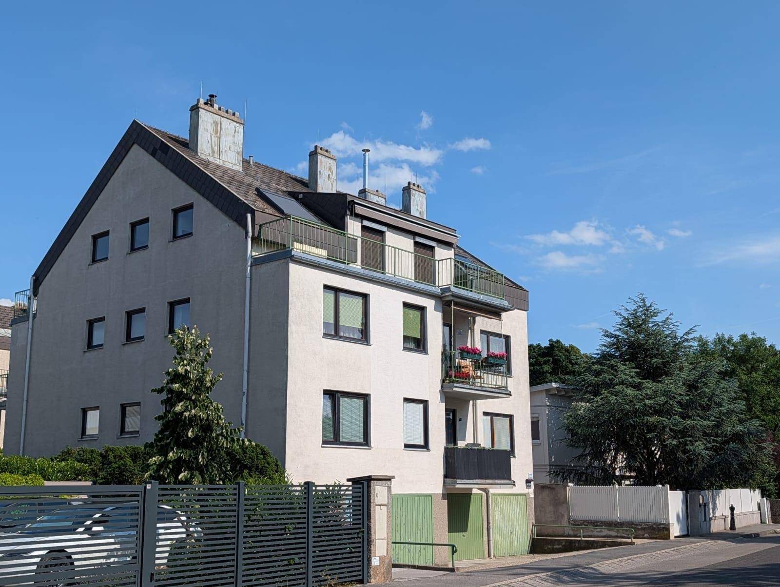 Mehrfamilienhaus mit mehreren Balkonen und grüner Umgebung unter blauem Himmel.