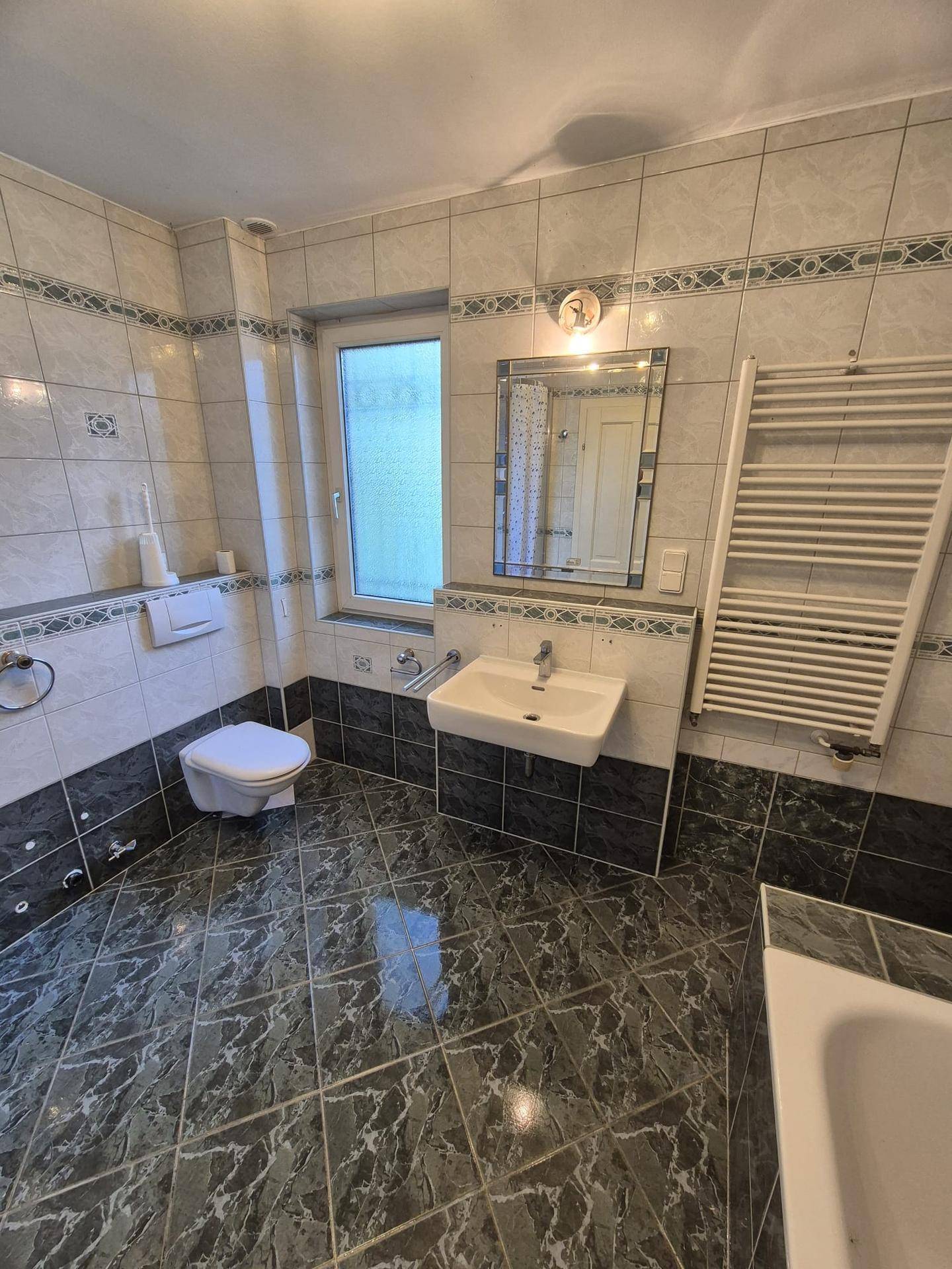 Geräumiges Badezimmer mit WC, Waschbecken, Spiegel und Fenster, komplett gefliest.
