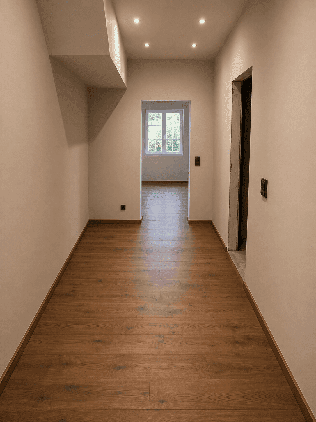 Heller Flur mit Holzboden, Einbaustrahlern und einem Fenster am Ende des Ganges.