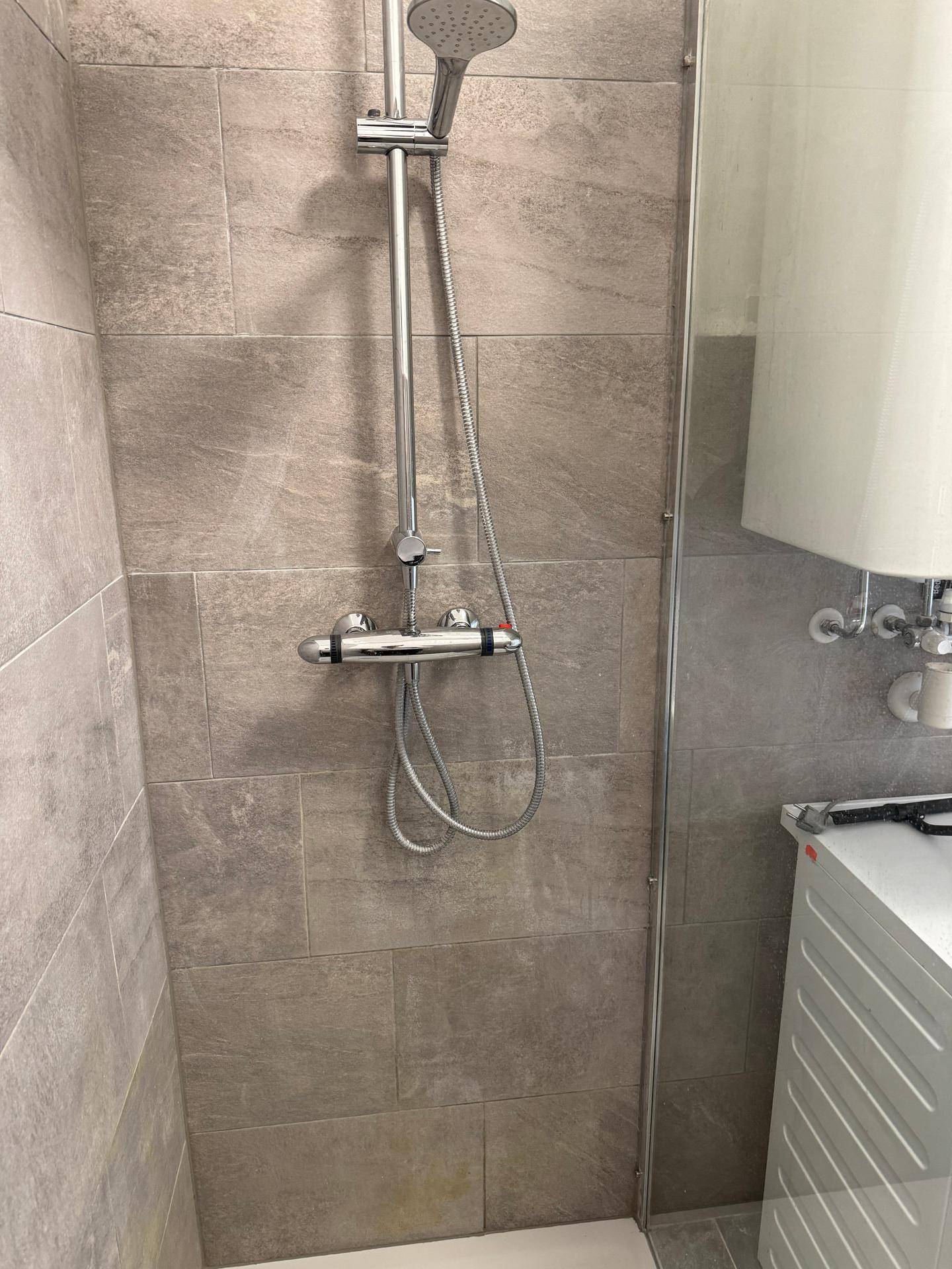 Duschbereich mit moderner Armatur und Handbrause, umgeben von grauen Fliesen und weißer Wand.
