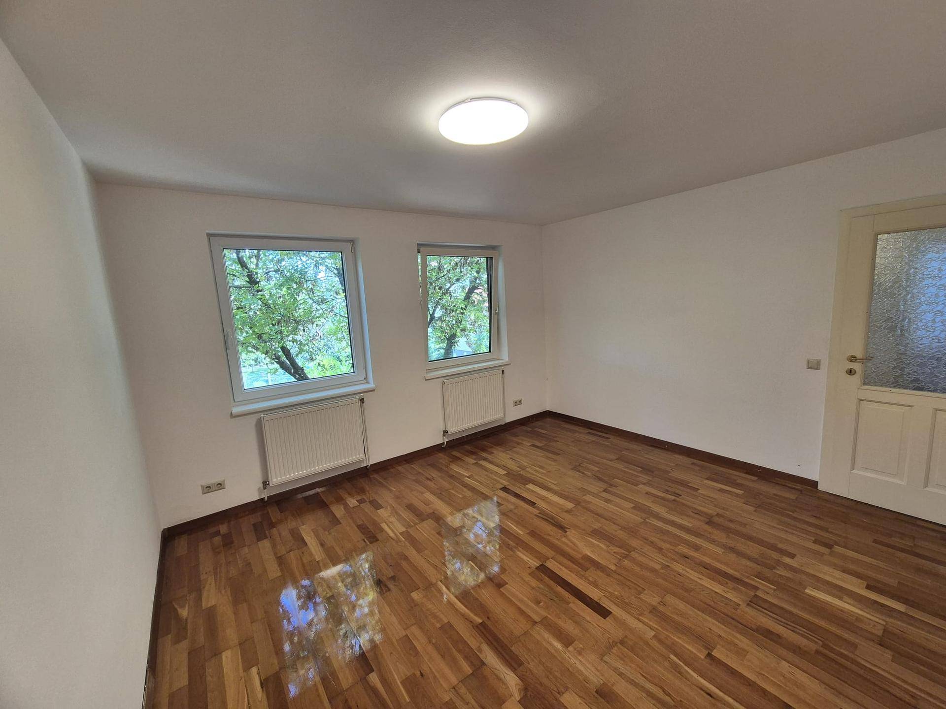 Helles Schlafzimmer mit glänzendem Parkettboden und zwei Fenstern mit Blick ins Grüne.