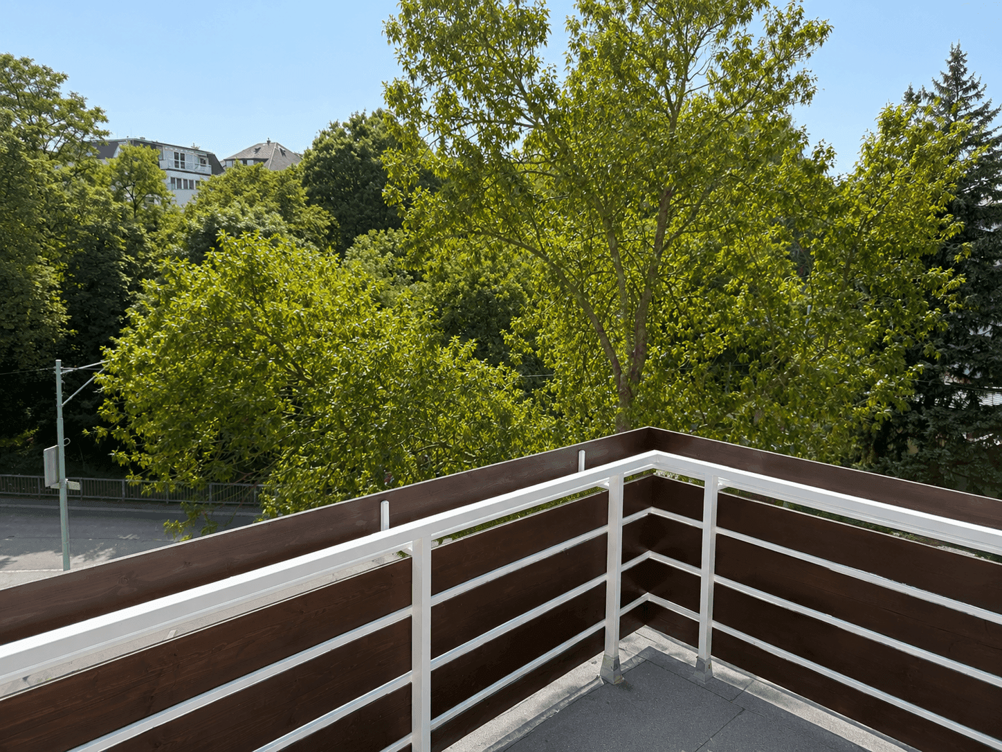 Blick von einem Balkon mit modernem Geländer auf grüne Bäume und die Straße.
