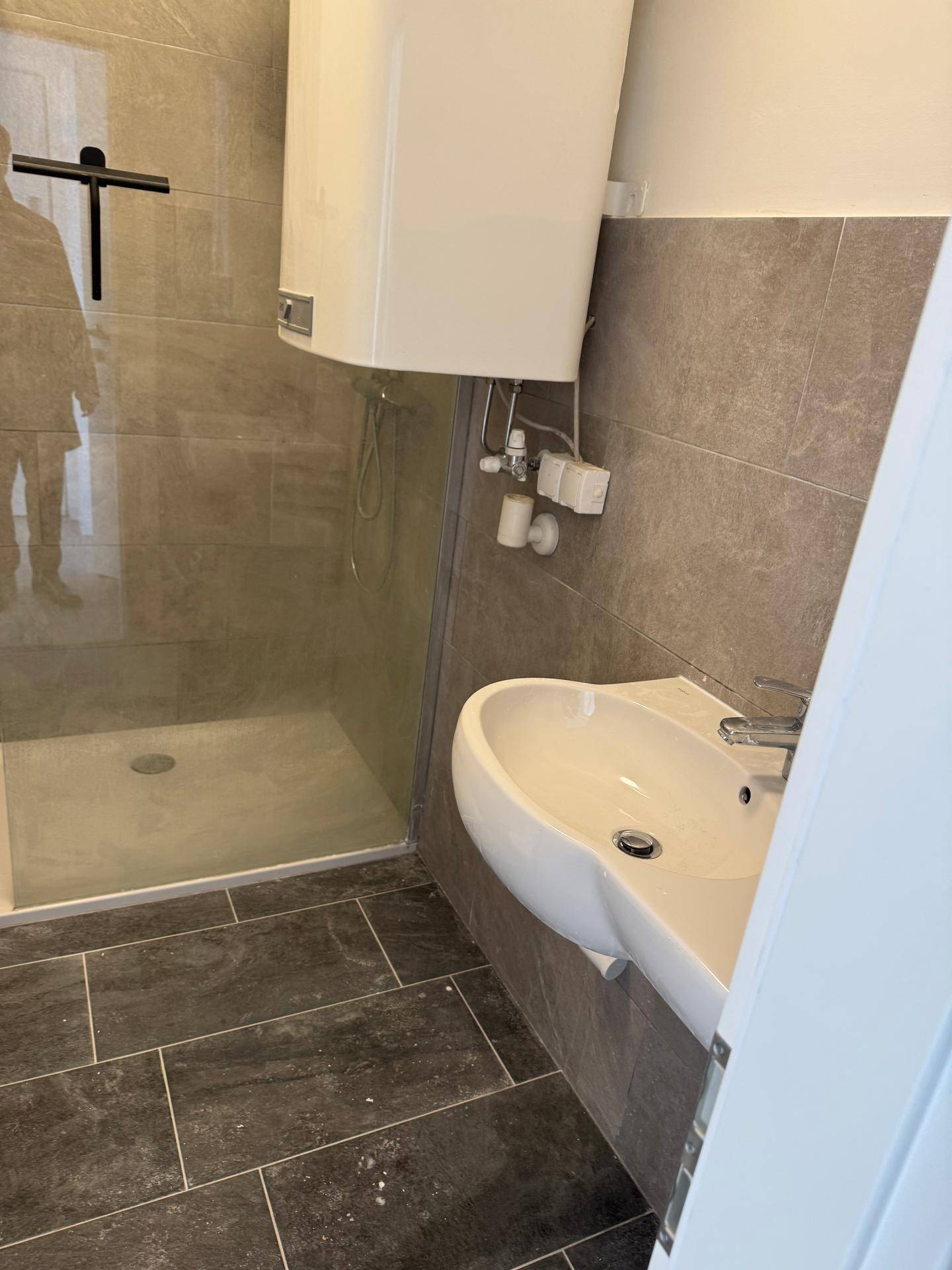 Badezimmer mit Wandwaschbecken, Dusche und Warmwasserboiler, in neutralen Grautönen gefliest.