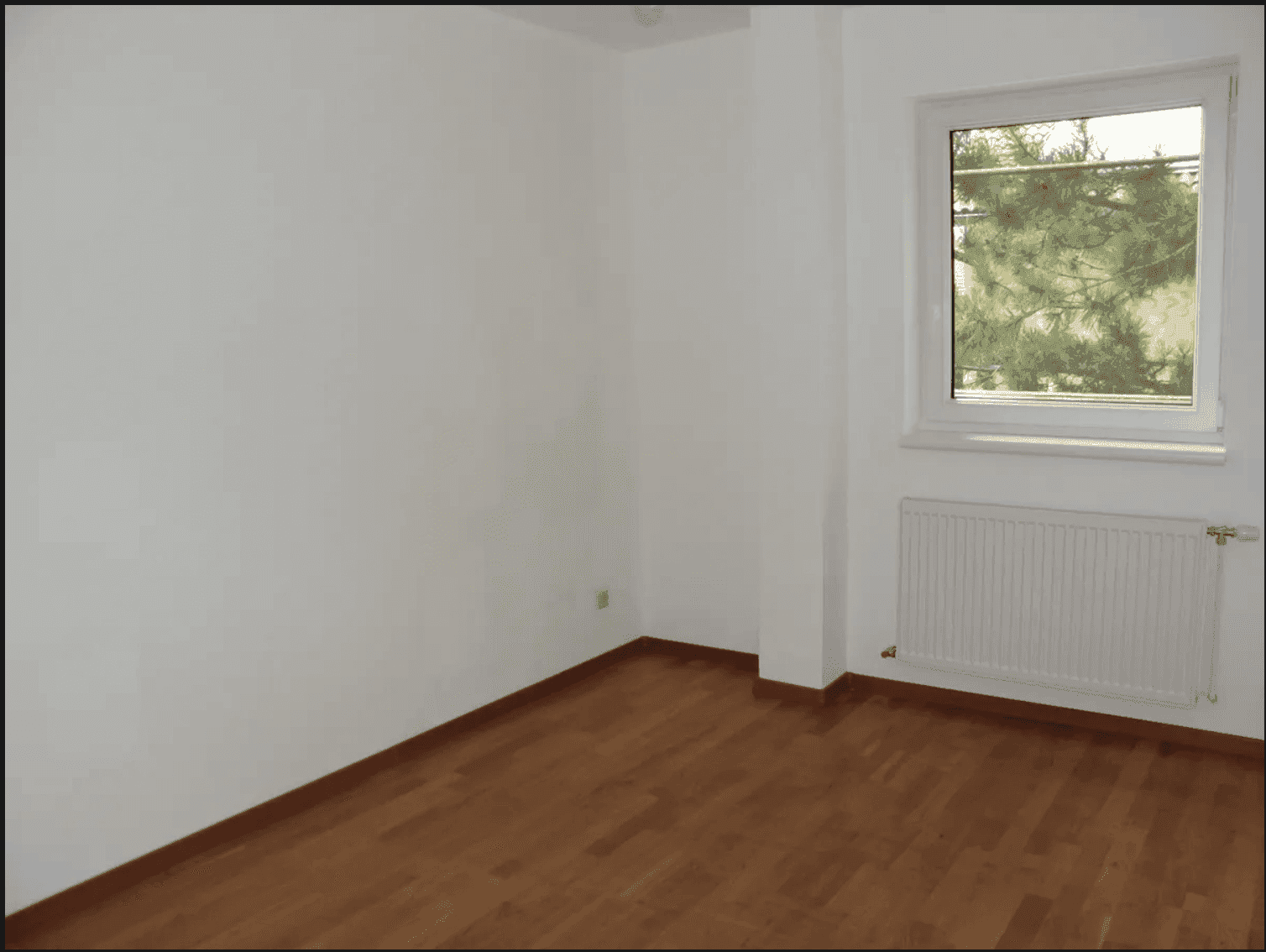 Ruhiges Zimmer mit Holzboden und einem Fenster, das viel Tageslicht hereinlässt und Grünblick bietet.