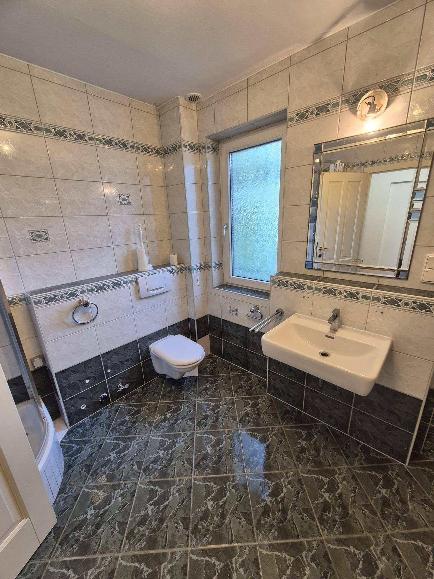 Gefliestes Badezimmer mit WC, Waschbecken, Spiegel und Fenster für natürliche Belüftung.