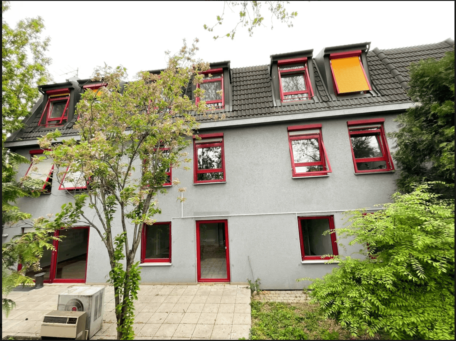Rückseite des Gebäudes mit grauer Fassade, roten Fensterrahmen und Dachgauben mit orangefarbenen Rollos.