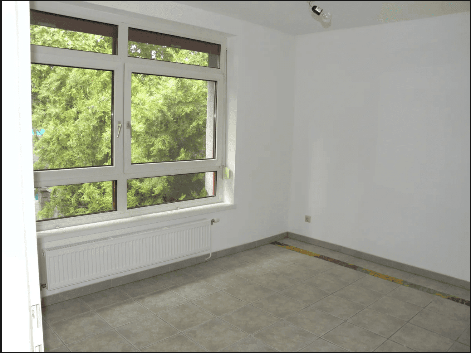 Geräumiger Raum mit Fliesenboden und einem großen Fenster, das viel Licht und Grünblick bietet.