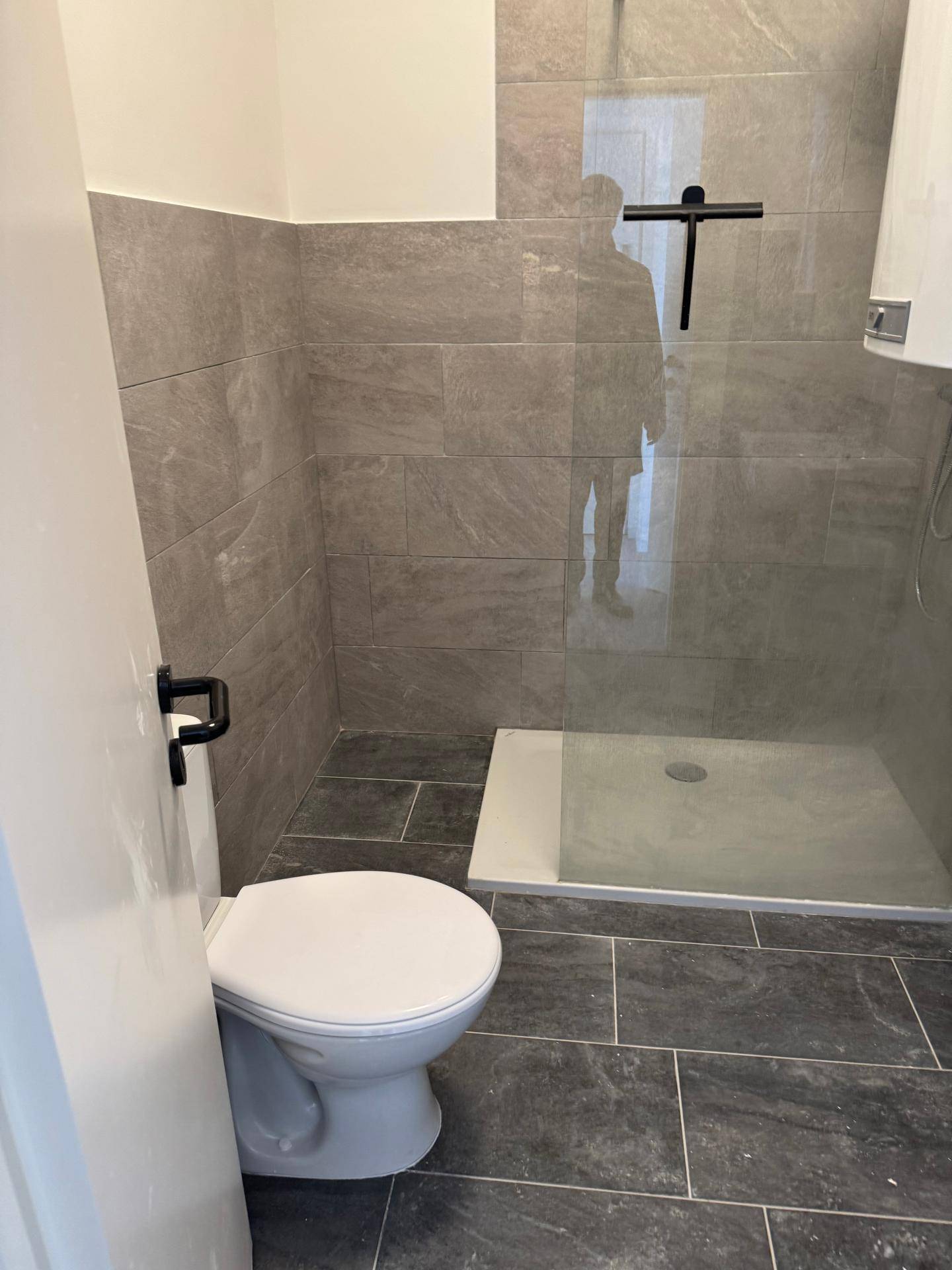 Modernes Badezimmer mit grauen Fliesen, Toilette und ebenerdiger Dusche mit Glasabtrennung.