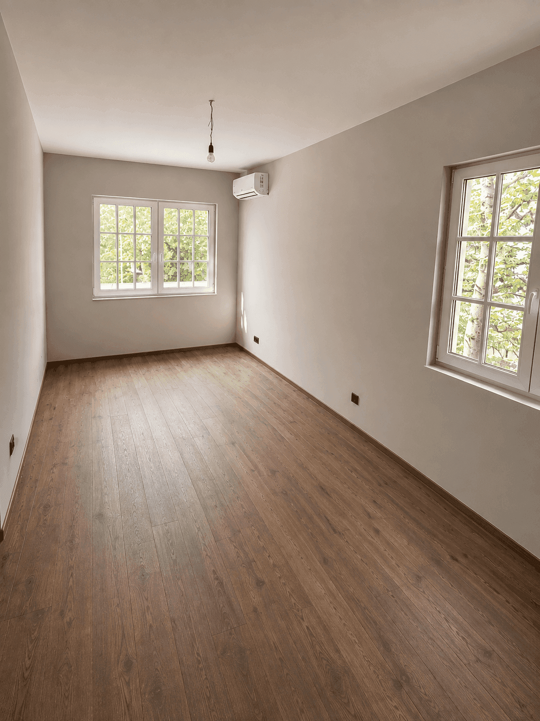 Helles Schlafzimmer mit Holzboden, zwei Fenstern und einer Klimaanlage.