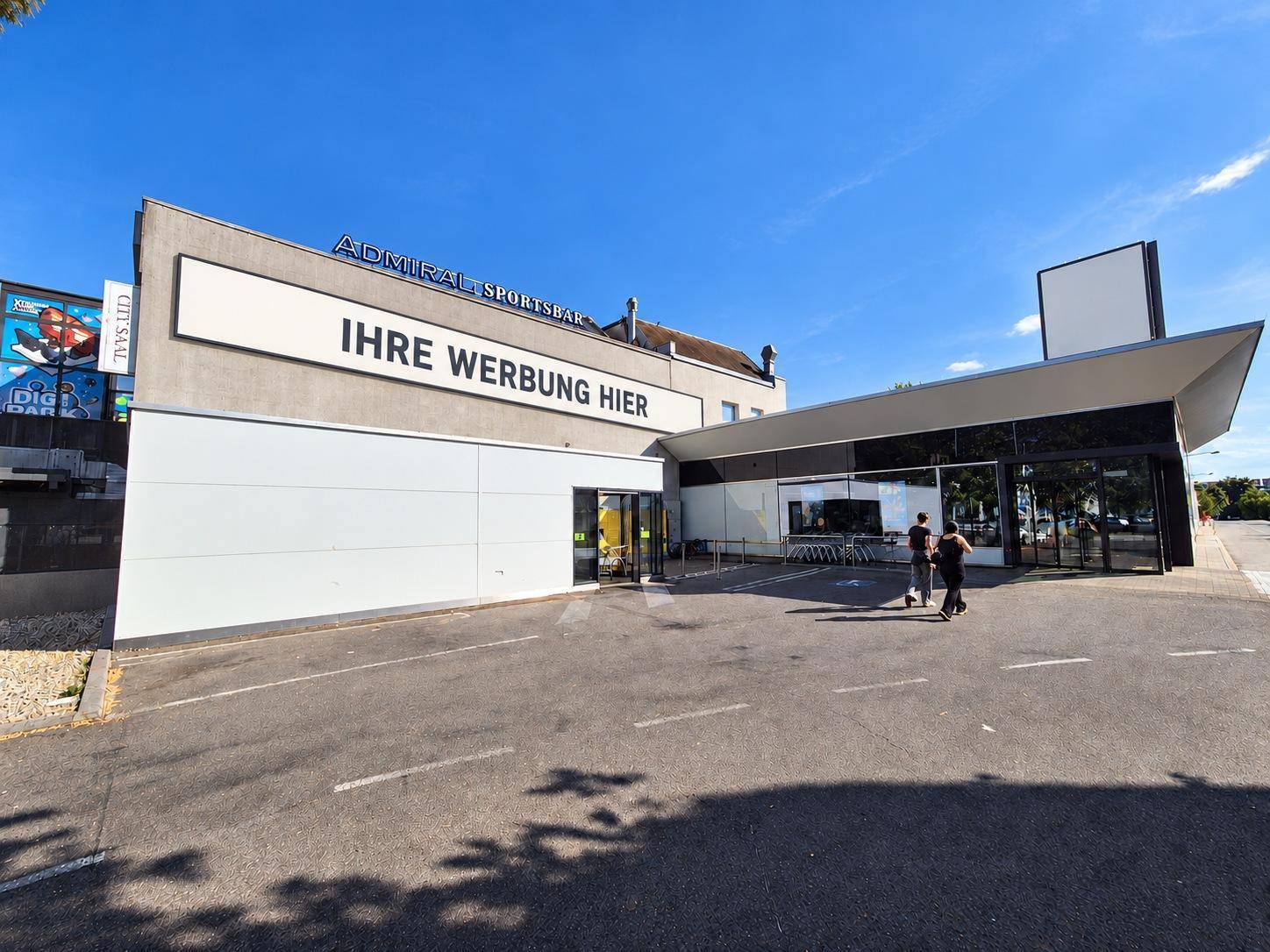 Weitläufige Außenansicht eines modernen Geschäftsgebäudes mit Werbefläche und Parkplatz, bei Tageslicht.