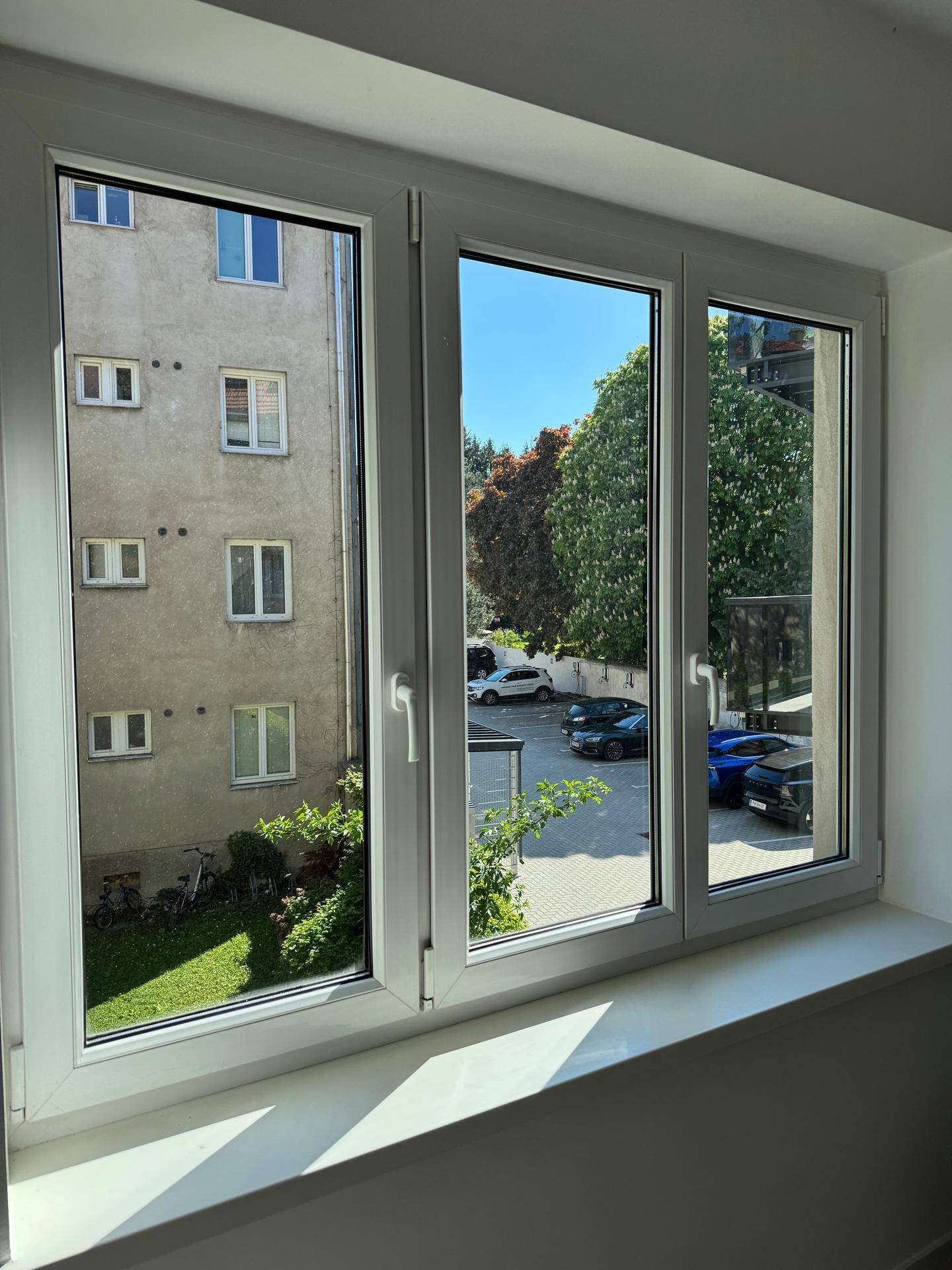 Fenster mit Blick auf einen begrünten Innenhof und parkende Autos an einem sonnigen Tag.