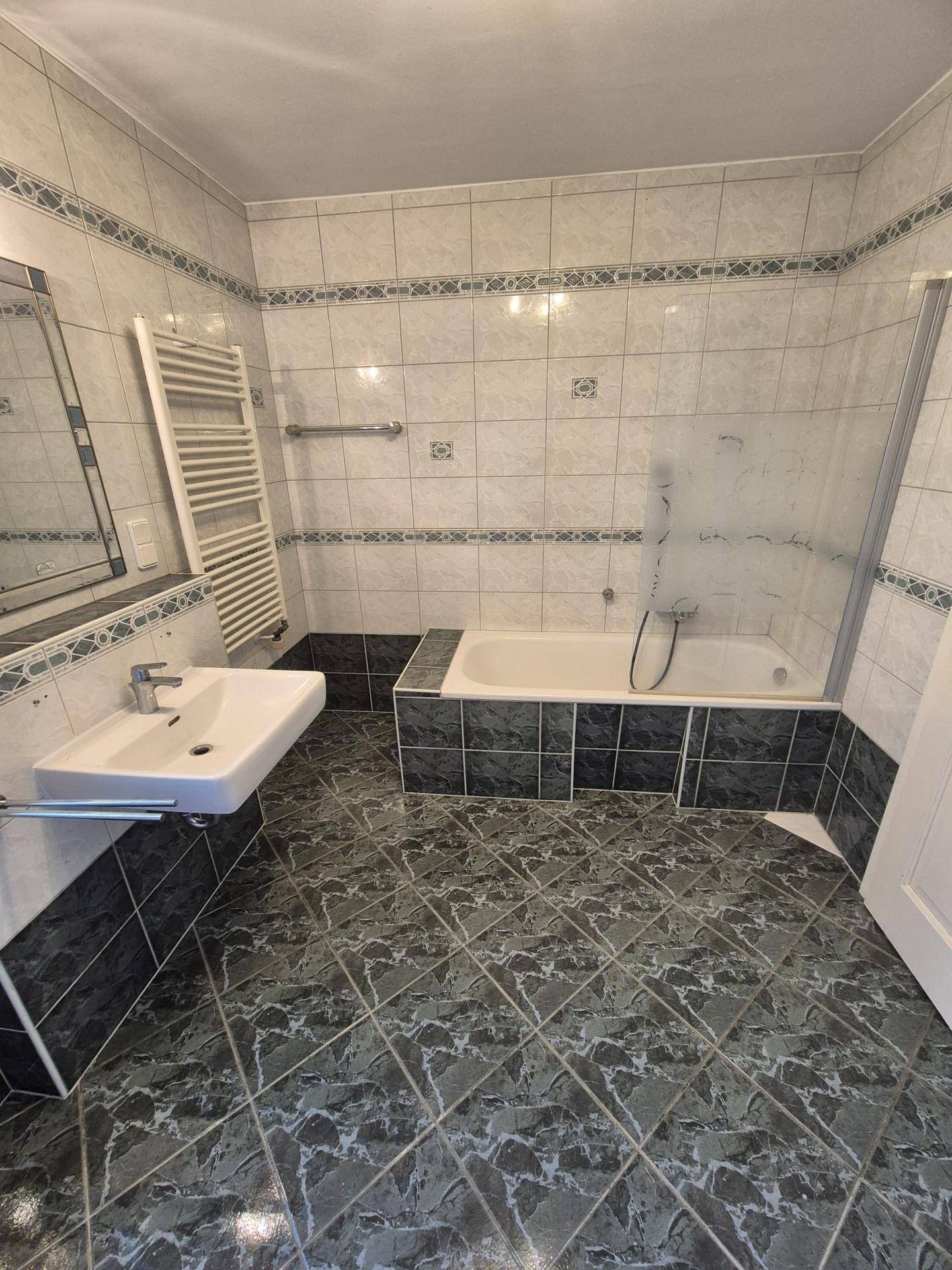 Gefliestes Badezimmer mit Badewanne, Duschabtrennung, Waschbecken und beheiztem Handtuchhalter.