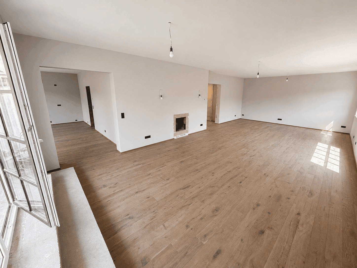 Weitläufiges Wohnzimmer mit hellem Holzboden, Kamin und viel Tageslicht durch große Fenster.