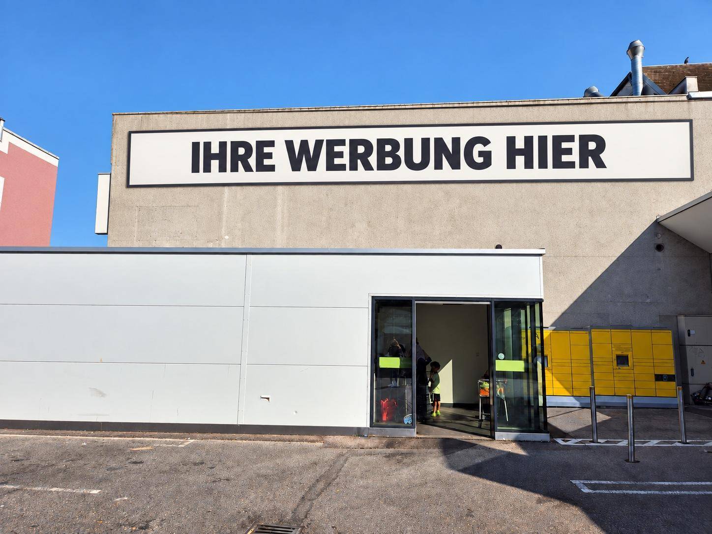 Eingangsbereich eines Geschäftsgebäudes mit großer Werbefläche und gelben Schließfächern im Freien.