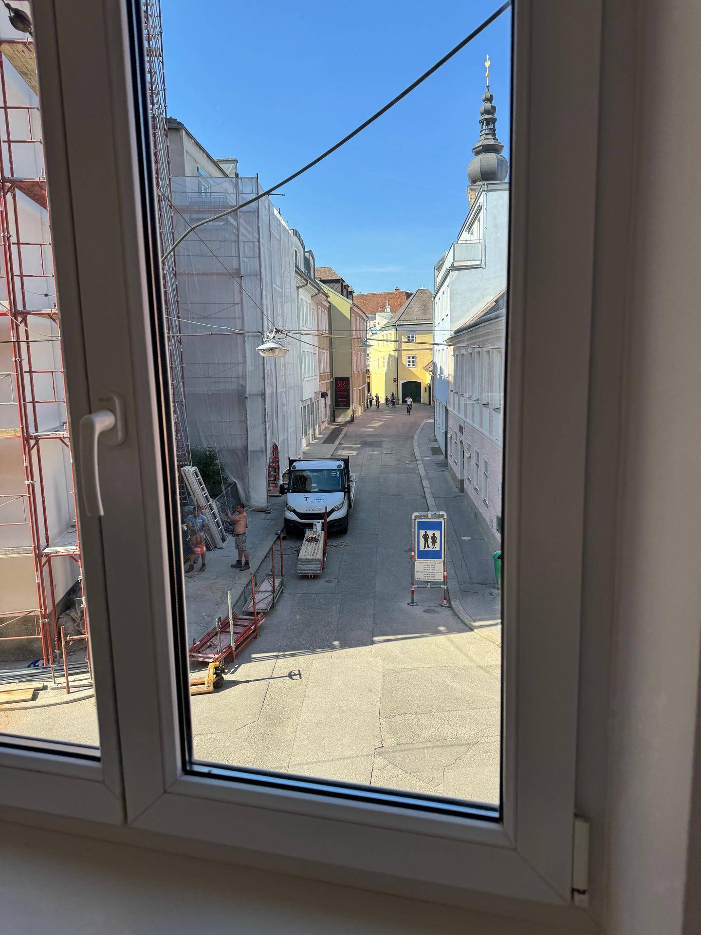 Fensterblick auf eine belebte Gasse mit Gerüsten und einem Kirchturm im sonnigen Hintergrund.