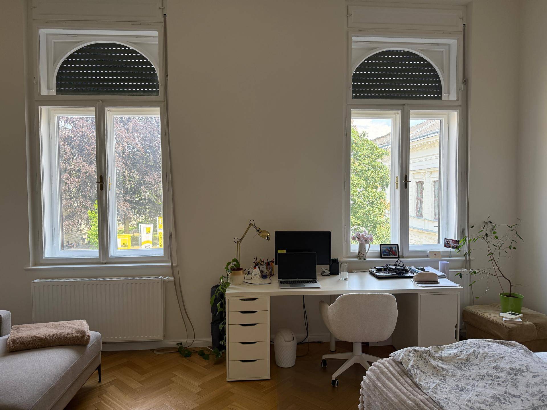 Heller Arbeitsbereich mit Schreibtisch, Computer und zwei hohen Fenstern mit Blick ins Grüne.