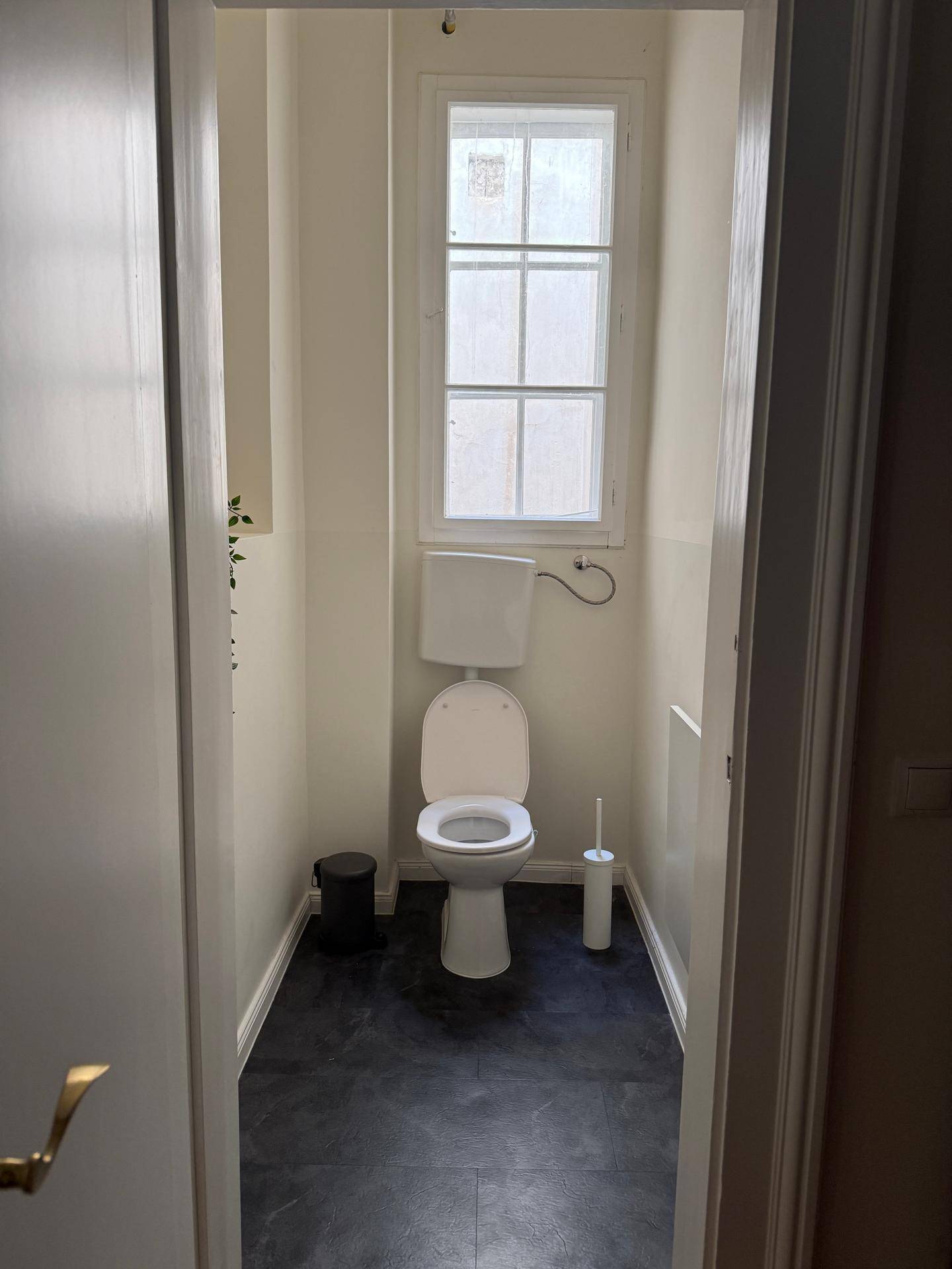 Schmale separate Toilette mit Fenster und dunklem Fliesenboden, sehr funktional.