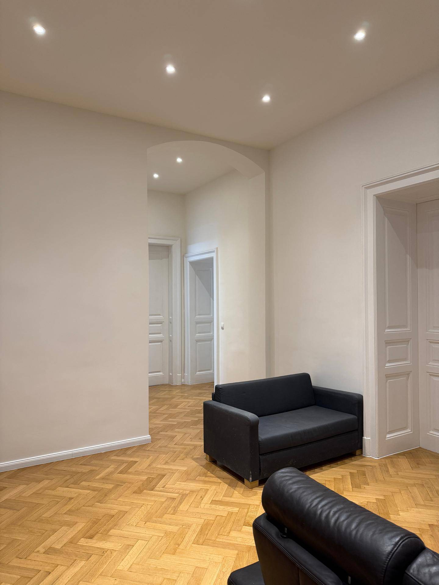 Heller Wohnbereich mit Fischgrätparkett, Einbaustrahlern und dunklen Sofas, modern eingerichtet.