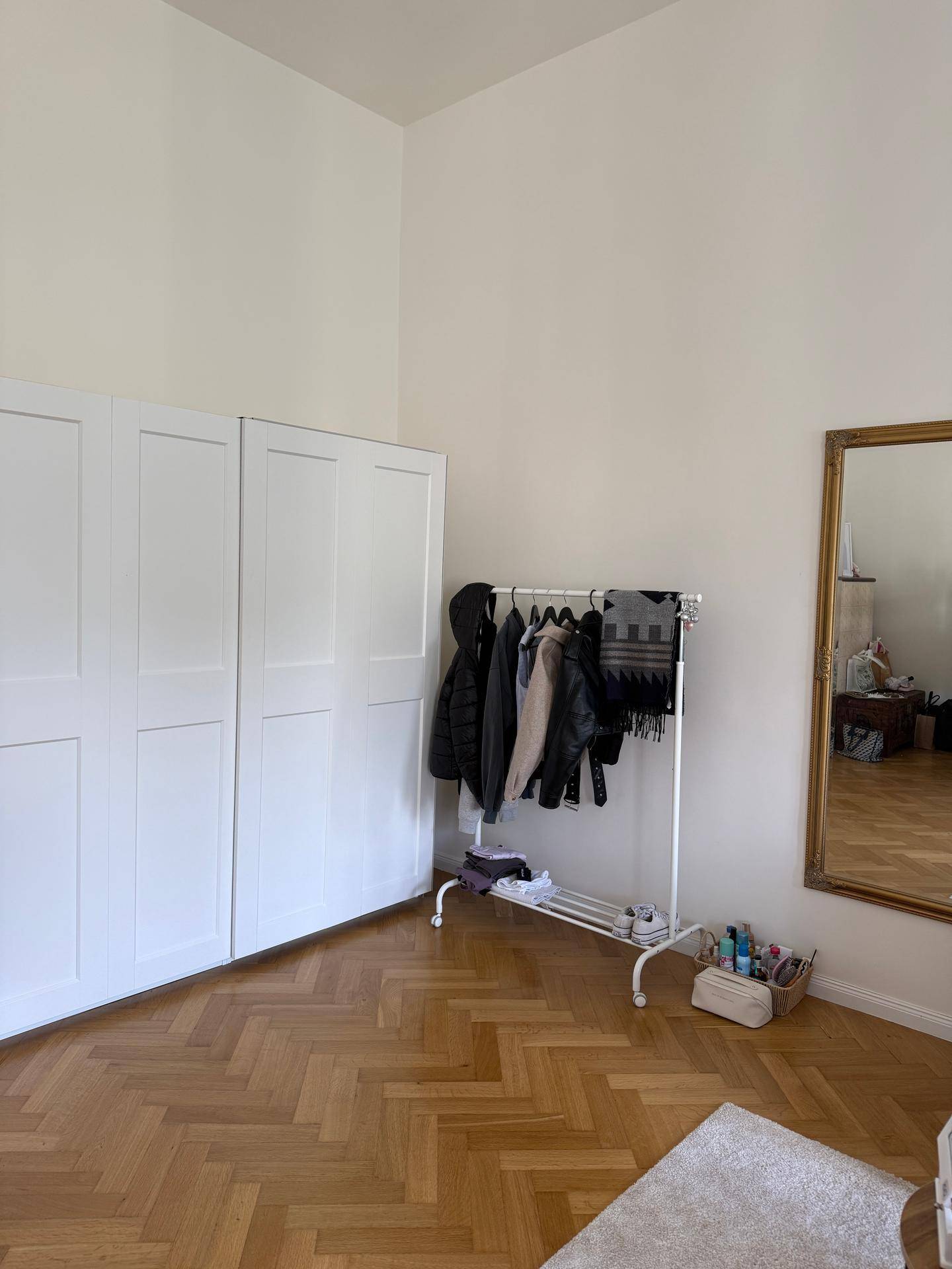 Heller Raum mit weißem Kleiderschrank, Kleiderstange und großem Spiegel, der viel Licht reflektiert.
