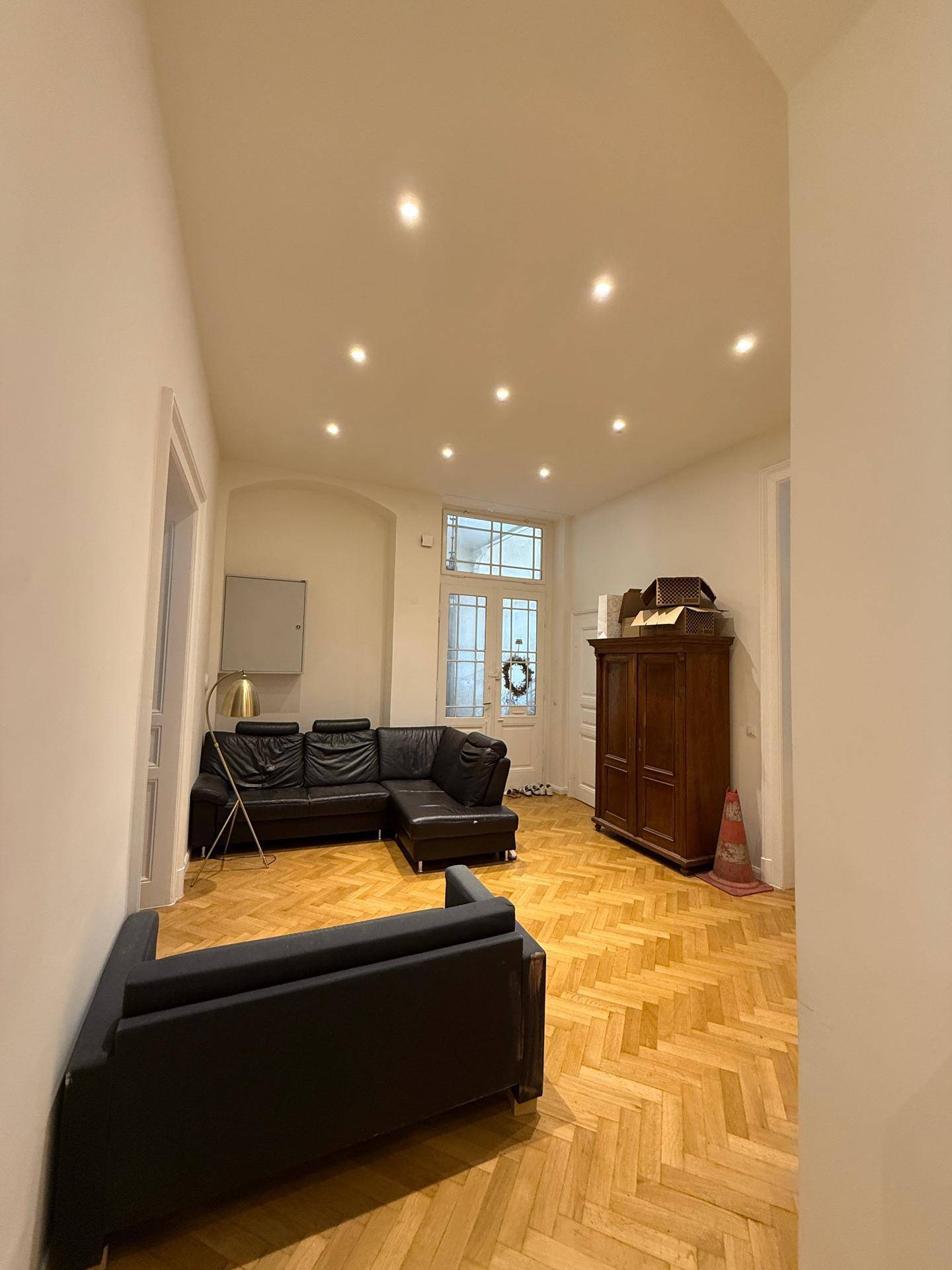 Geräumiges Wohnzimmer mit Fischgrätparkett, dunklem Sofa und antikem Holzschrank, elegant gestaltet.