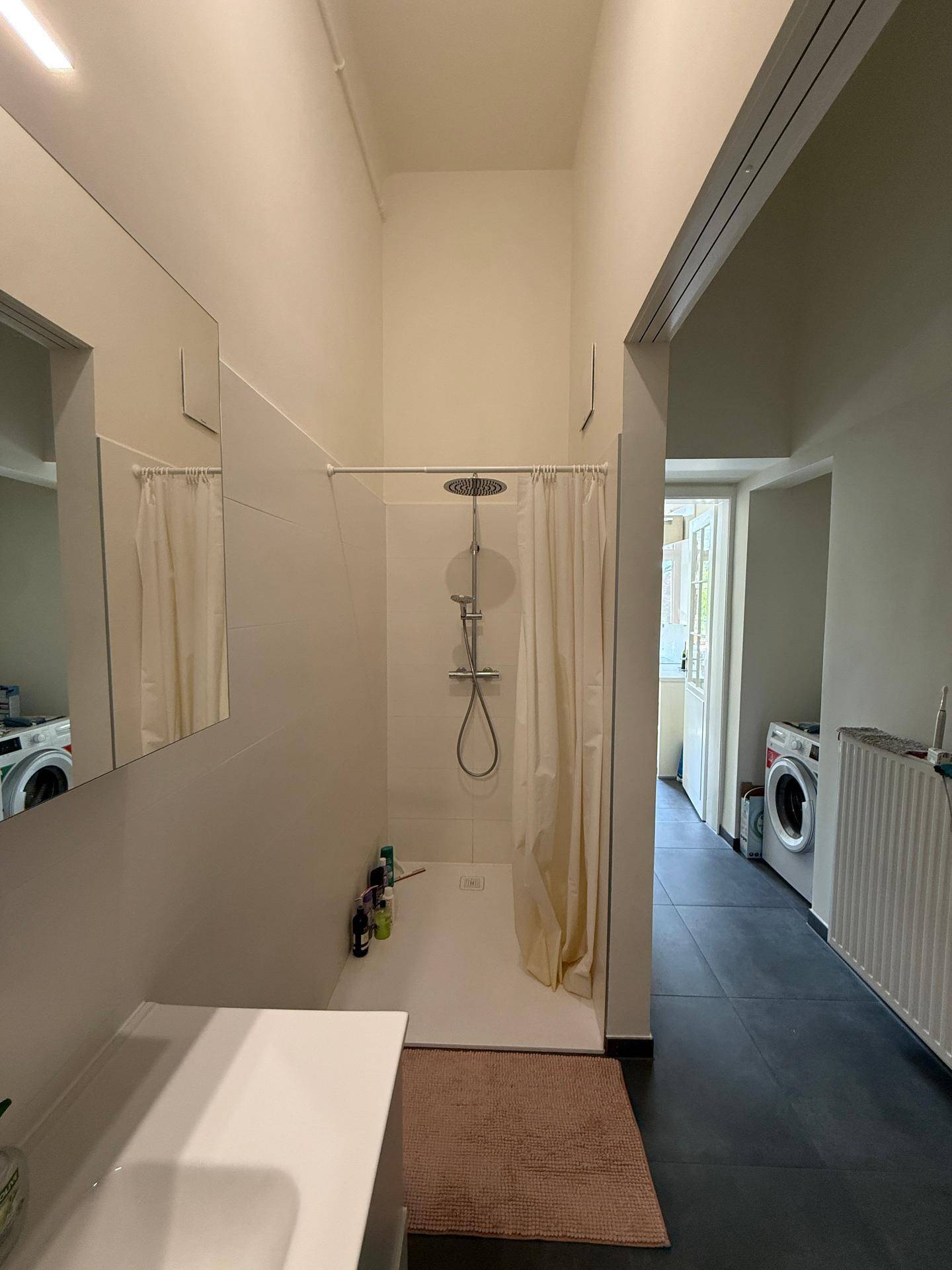 Geräumiges Badezimmer mit ebenerdiger Dusche und Blick auf den Waschmaschinenbereich im Hintergrund.