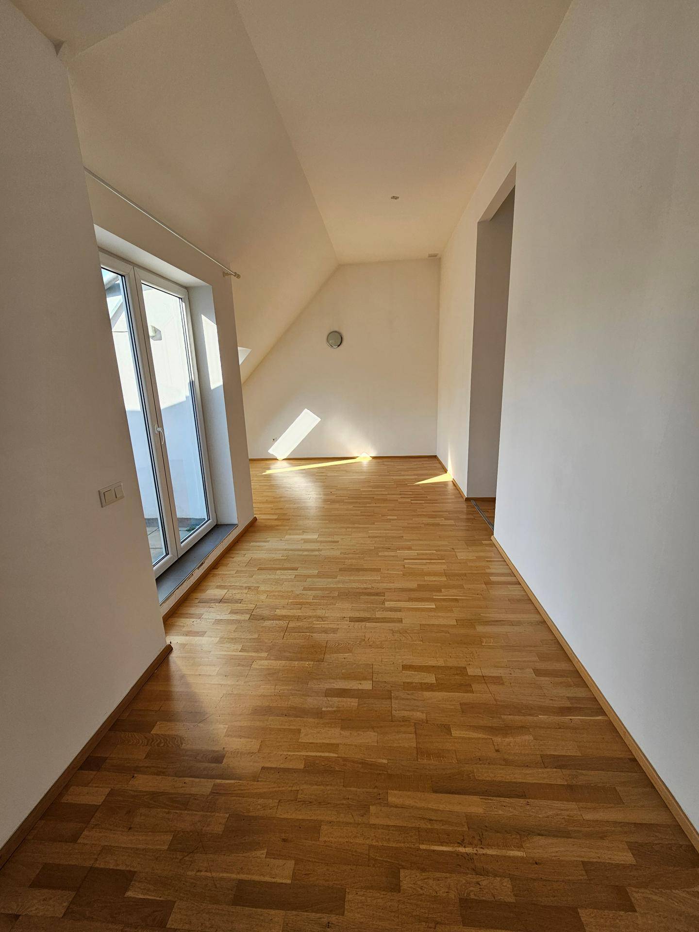 Heller Raum mit Holzboden, Dachschräge und Zugang zu einer Terrasse, durchflutet von Sonnenlicht.