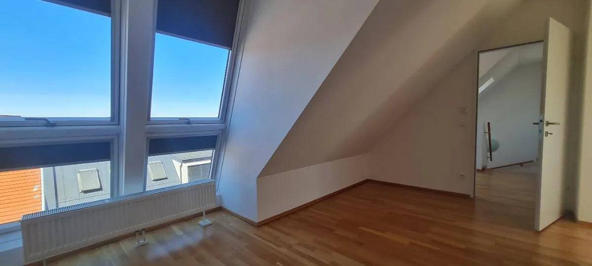 Heller Raum mit Holzboden, Dachschräge und großen Fenstern, die viel Tageslicht hereinlassen.