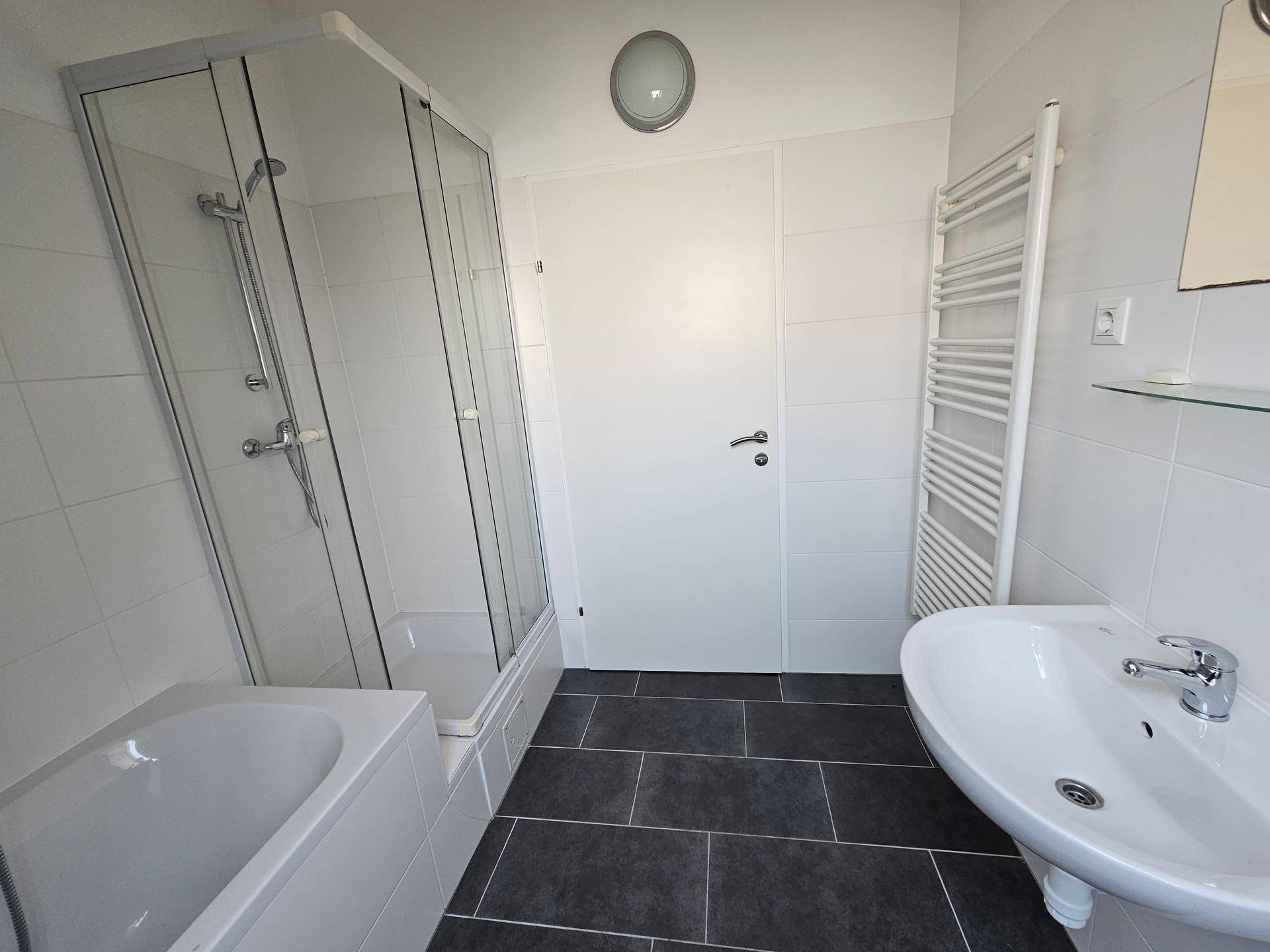 Helles Badezimmer mit Duschkabine, Waschbecken, beheiztem Handtuchhalter und dunklen Fliesen.