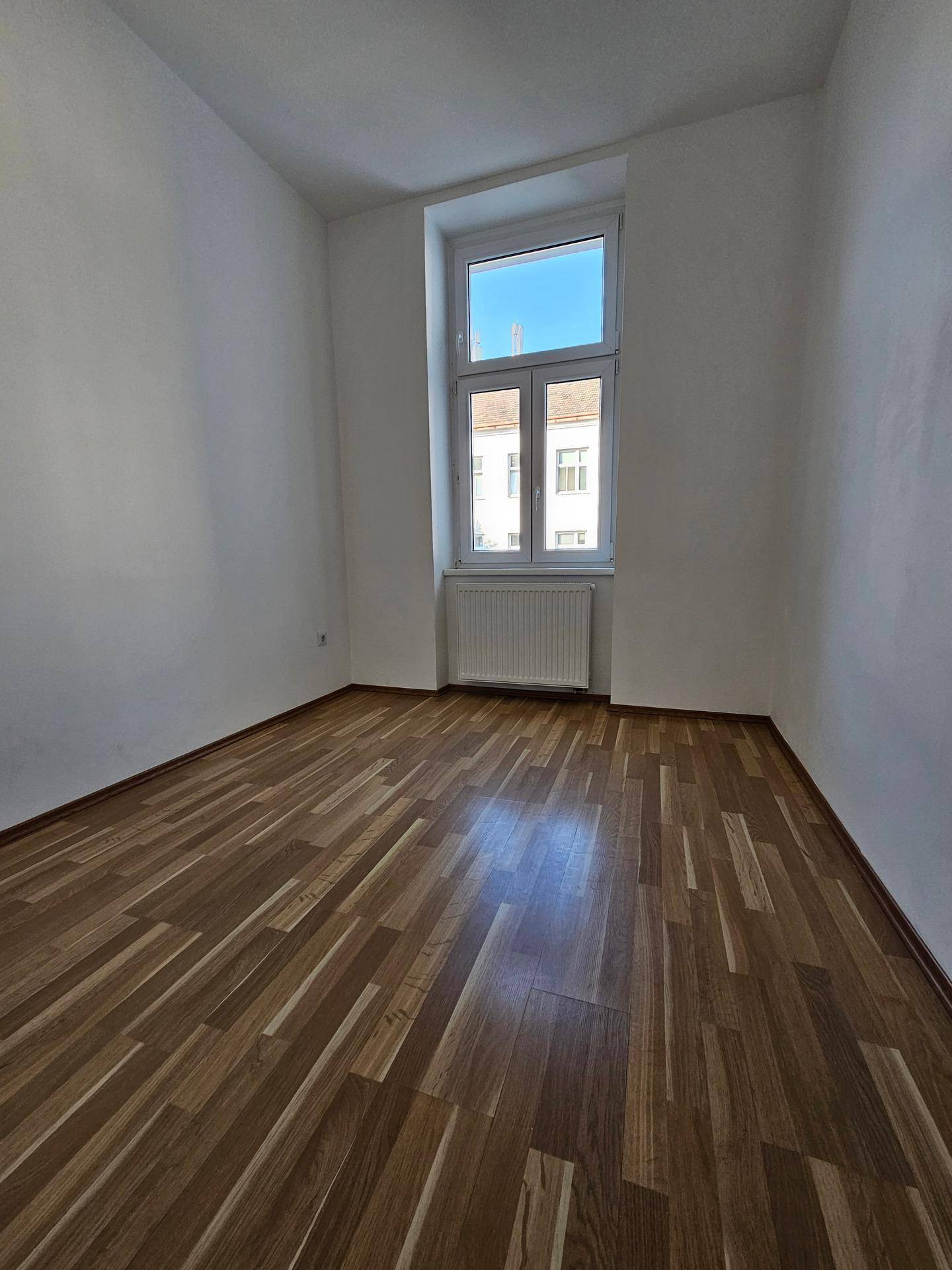 Heller Raum mit Holzparkettboden und einem großen Fenster, das viel Tageslicht hereinlässt.