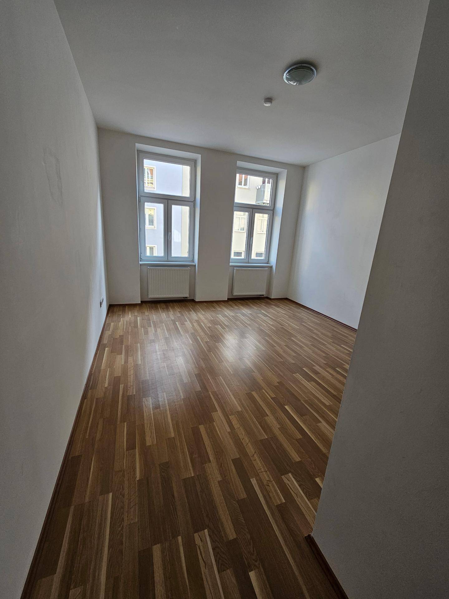 Heller Raum mit zwei Fenstern und warmem Holzparkettboden, ideal als Schlaf- oder Wohnzimmer.