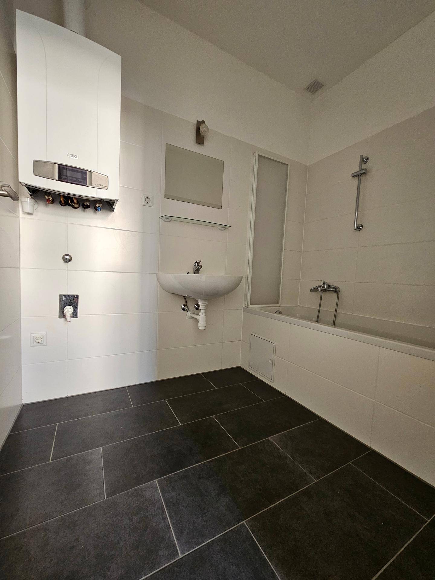 Badezimmer mit Badewanne, Waschbecken und Gastherme an der Wand, gefliest in Weiß und Anthrazit.