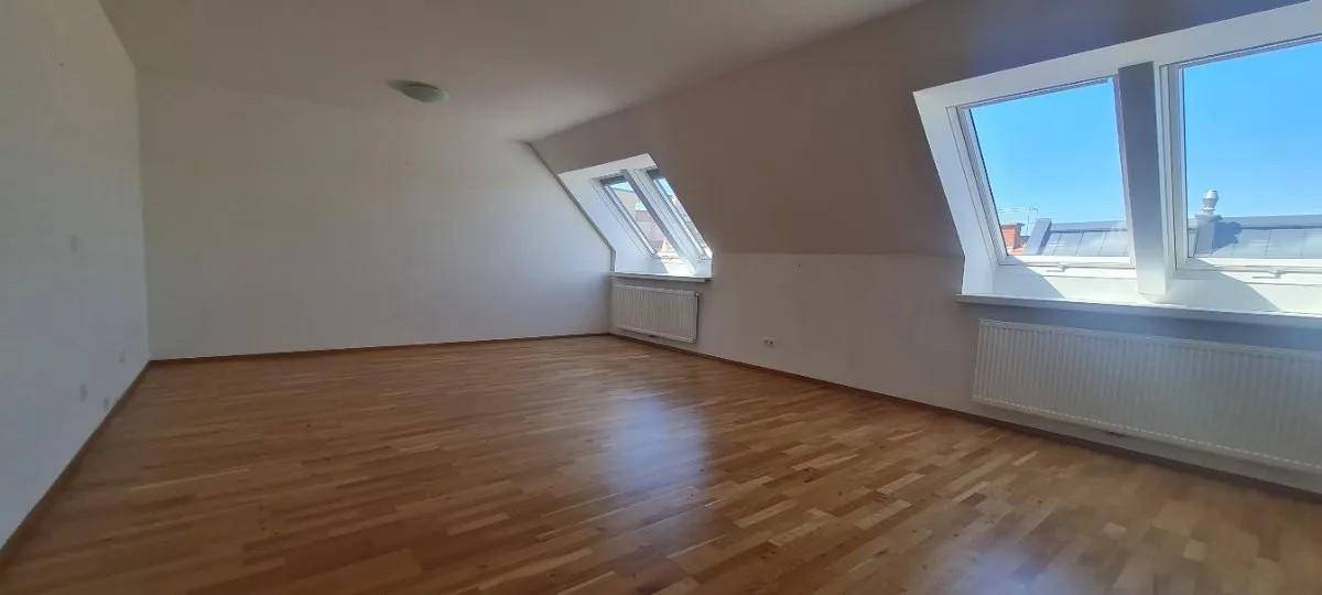 Geräumiger Raum mit Holzboden, Dachschräge und mehreren Fenstern, die für eine helle Atmosphäre sorgen.