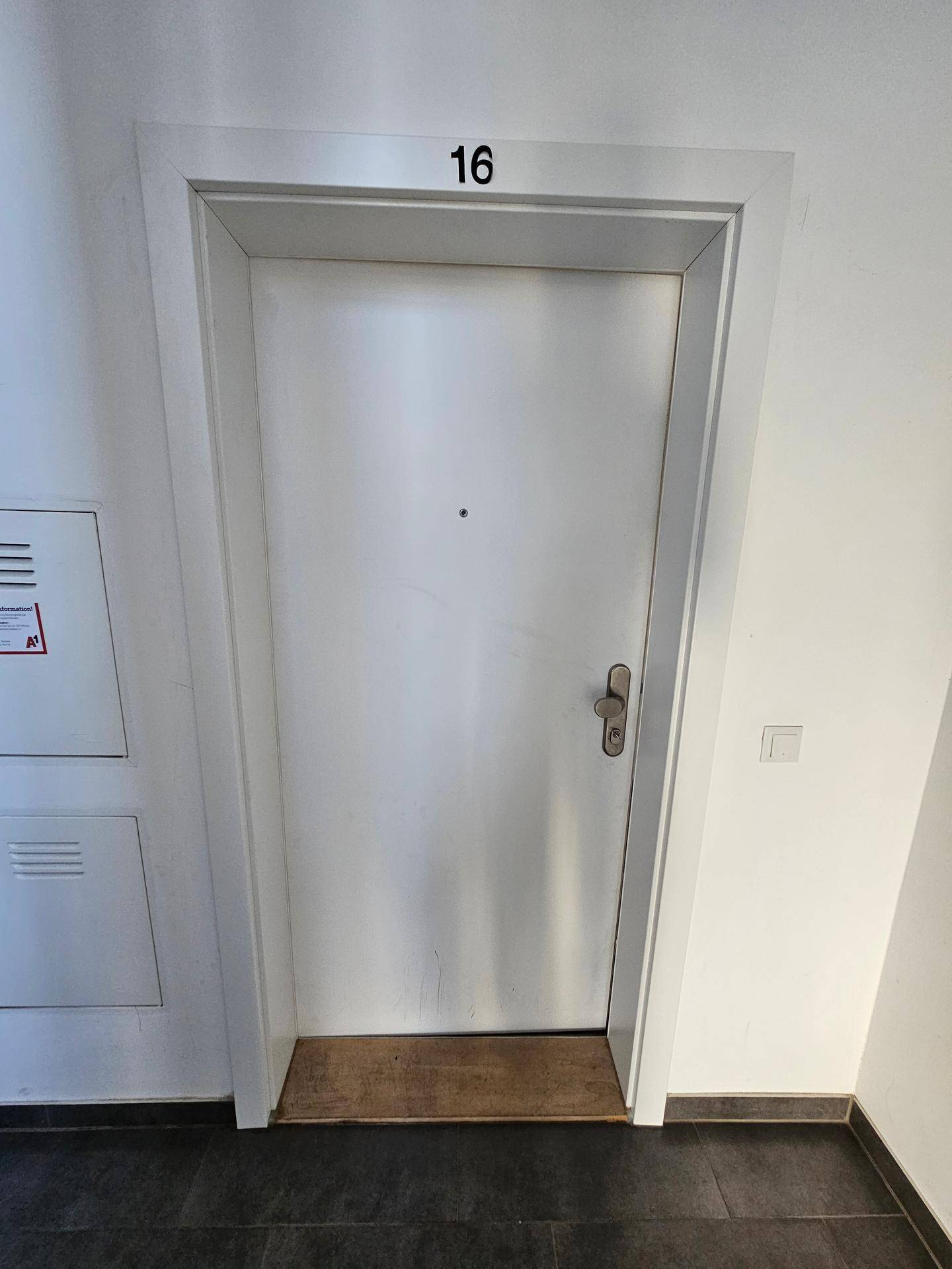 Eingangstür der Wohnung Nummer 16 mit dunklem Fliesenboden und weißem Rahmen.