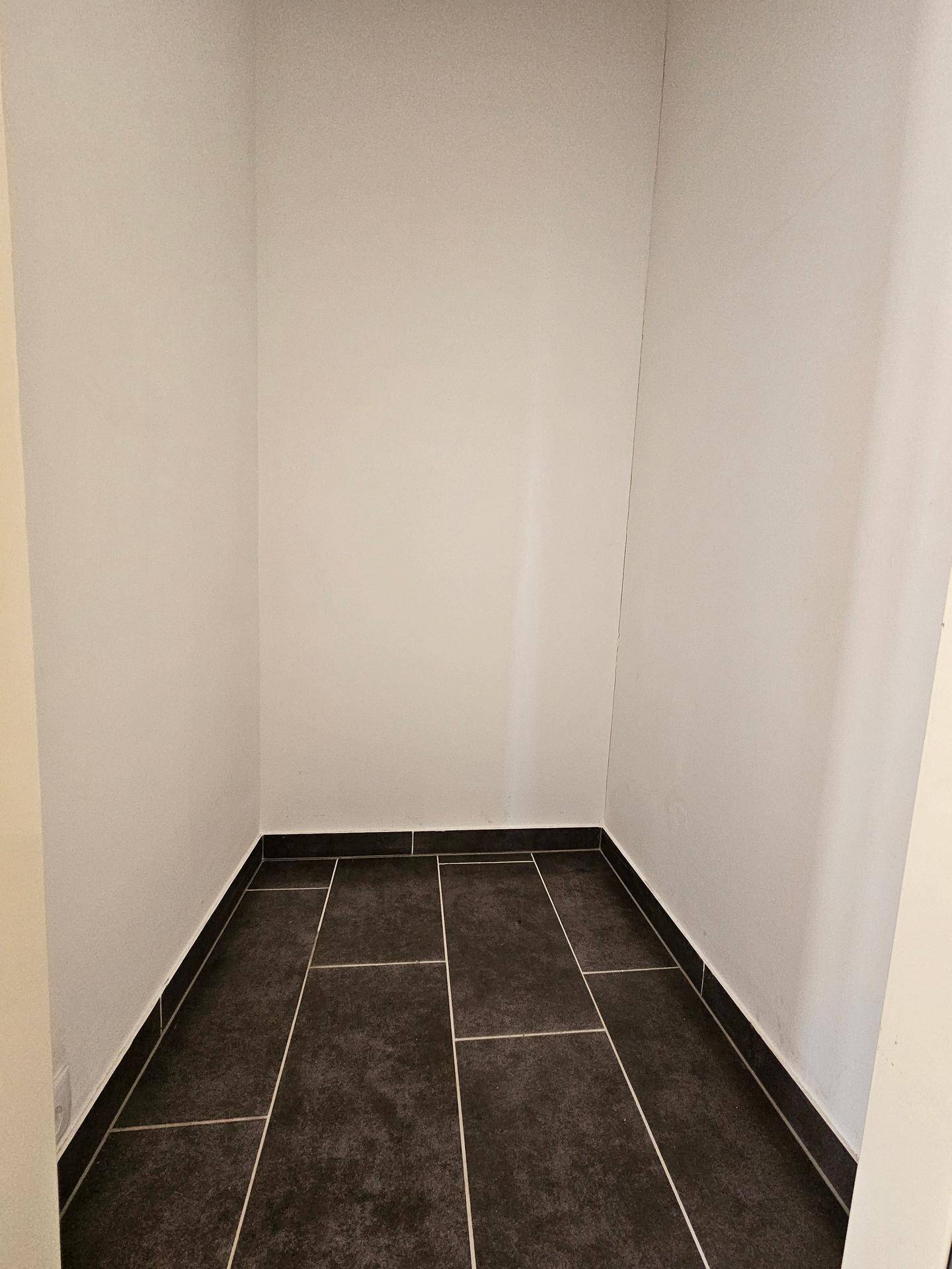 Leerer Abstellraum mit dunklem Fliesenboden und weißen Wänden, ideal für Lagerzwecke.