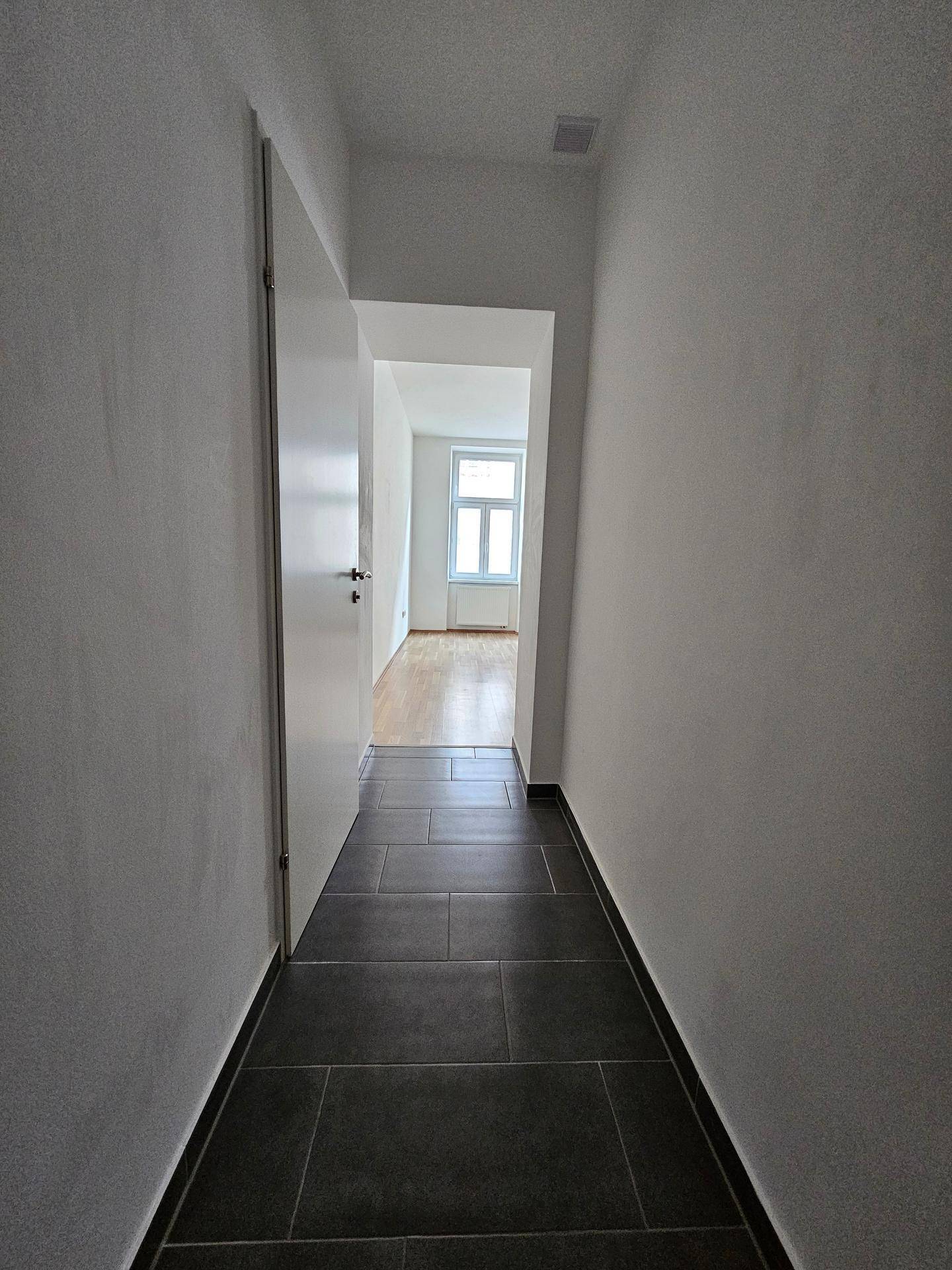 Langer Flur mit dunklen Fliesen, der den Blick auf einen hellen Raum mit Holzfußboden freigibt.
