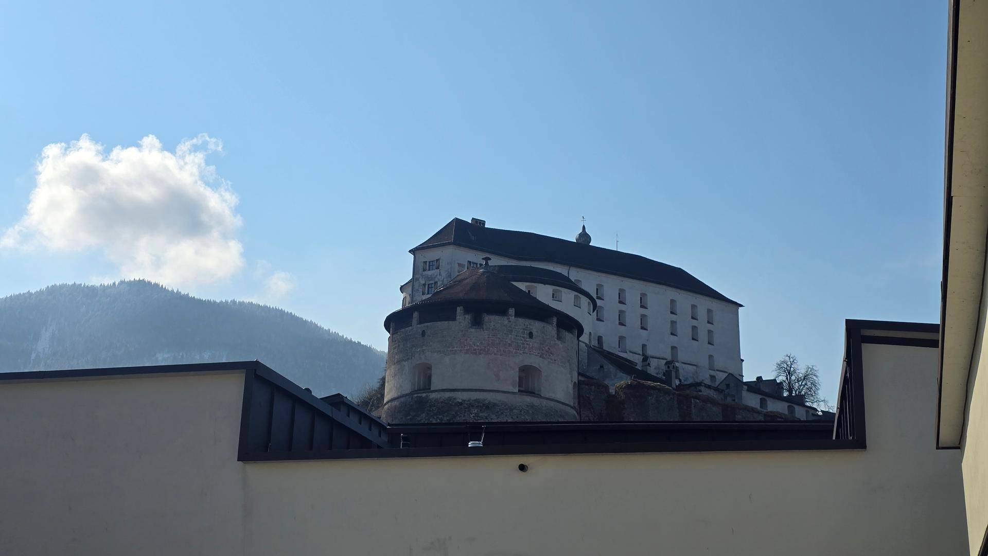 Historische Burg auf einem Hügel mit Bergen im Hintergrund unter blauem Himmel.