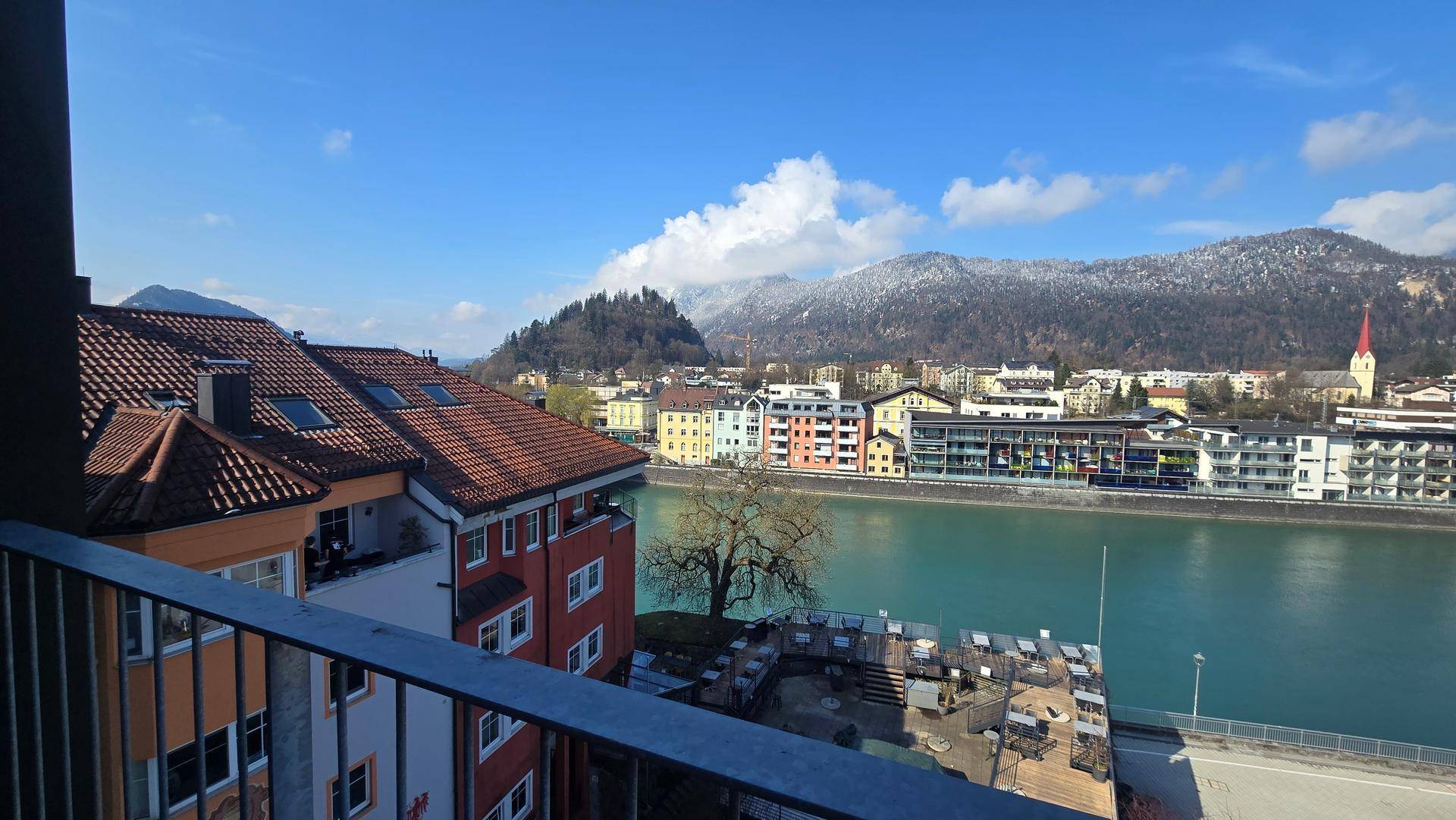 Balkonblick auf eine malerische Stadt mit Fluss und schneebedeckten Bergen im Hintergrund.