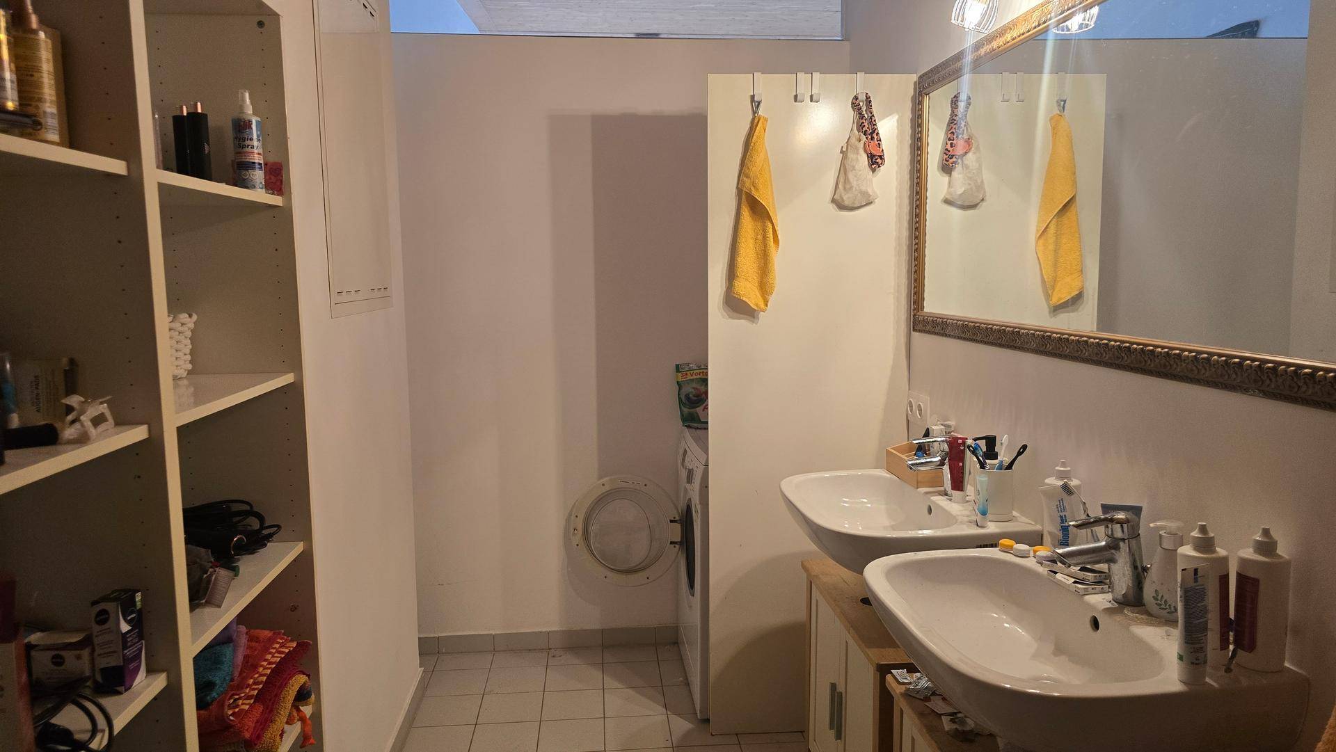 Badezimmer mit Doppelwaschbecken, Spiegel, Regalen und einer Waschmaschine im Hintergrund.