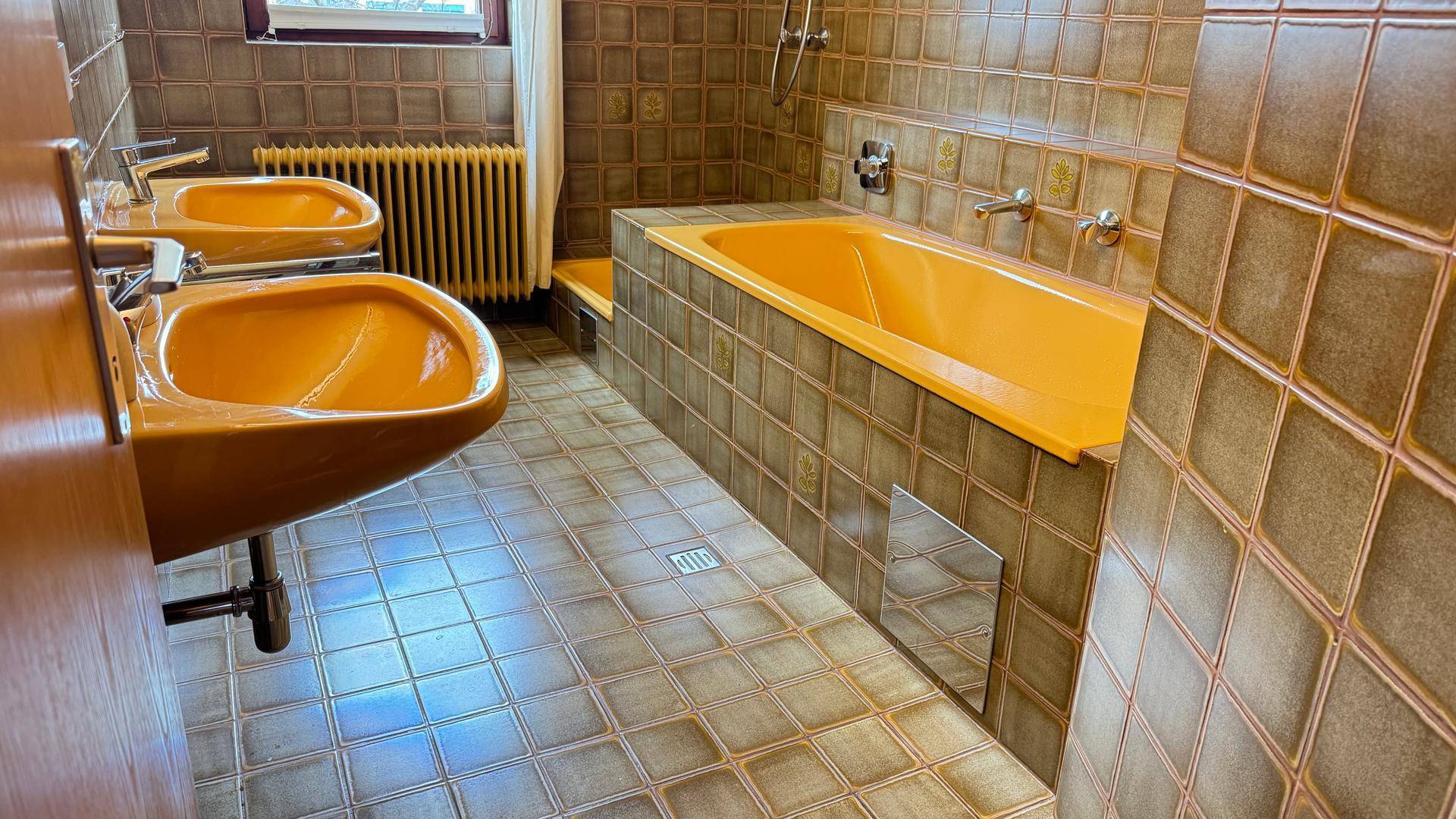 Badezimmer mit gelber Badewanne, Doppelwaschbecken und braunen Fliesen im 80er-Jahre-Stil.