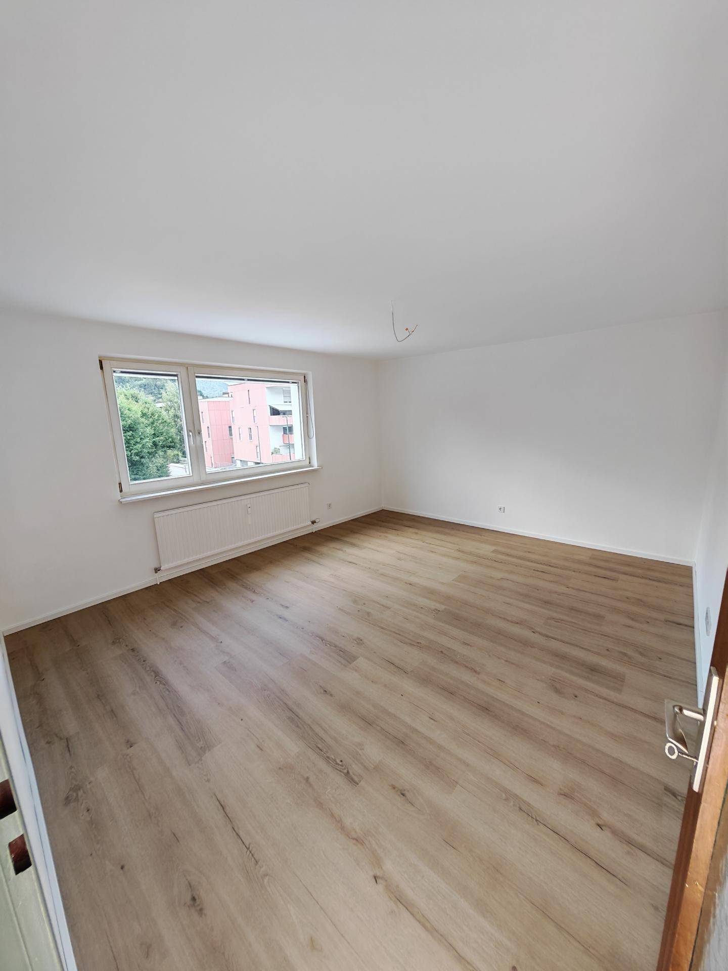 Helles, leeres Zimmer mit Laminatboden und Fenster mit Blick auf die Nachbargebäude.