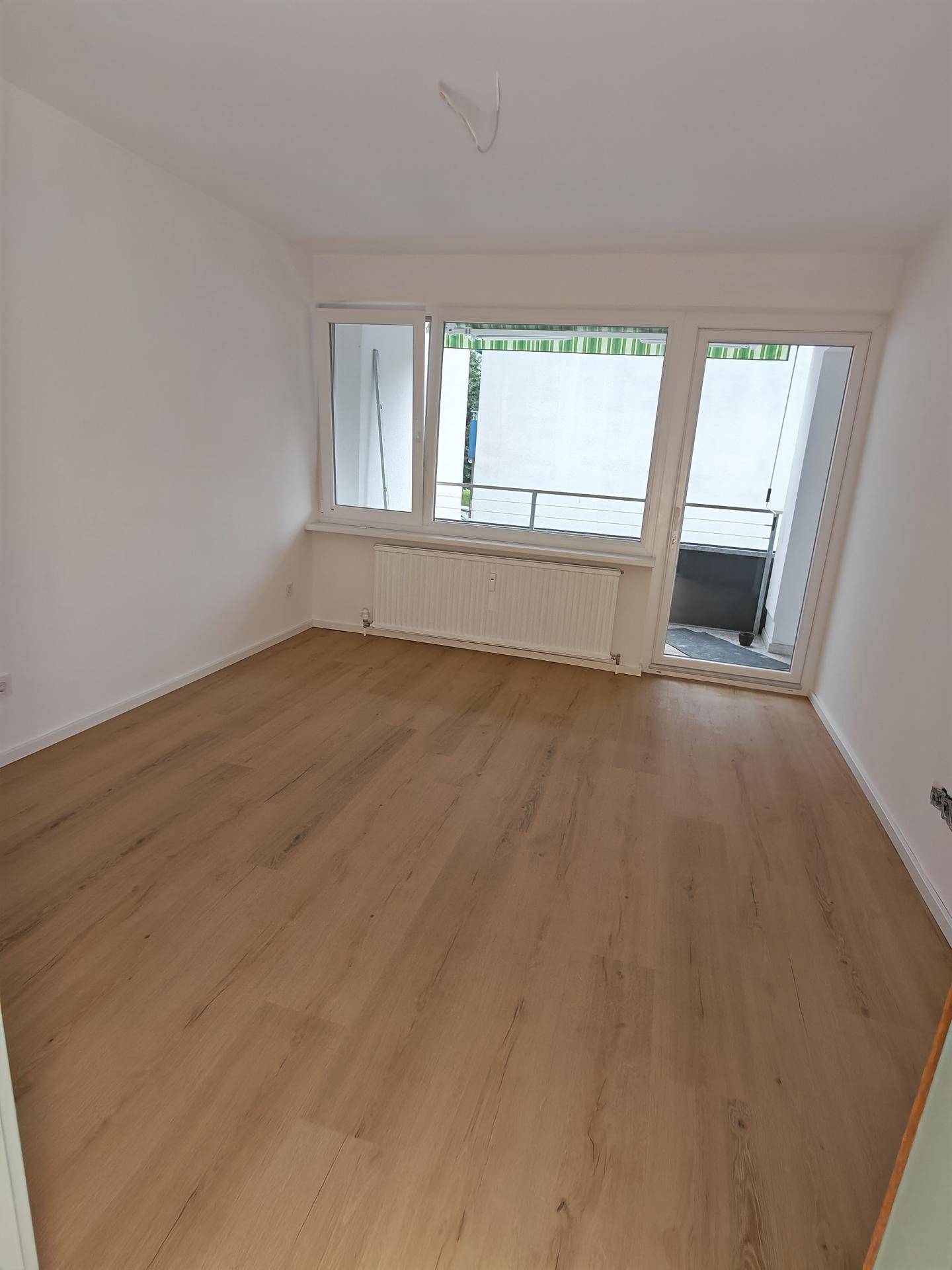 Leeres Zimmer mit hellem Laminatboden, weißen Wänden und Balkonzugang.