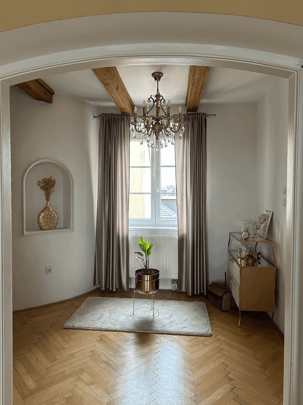 Helles Zimmer mit Fischgrätparkett, sichtbaren Holzbalken und einem eleganten Kronleuchter über einem Teppich.