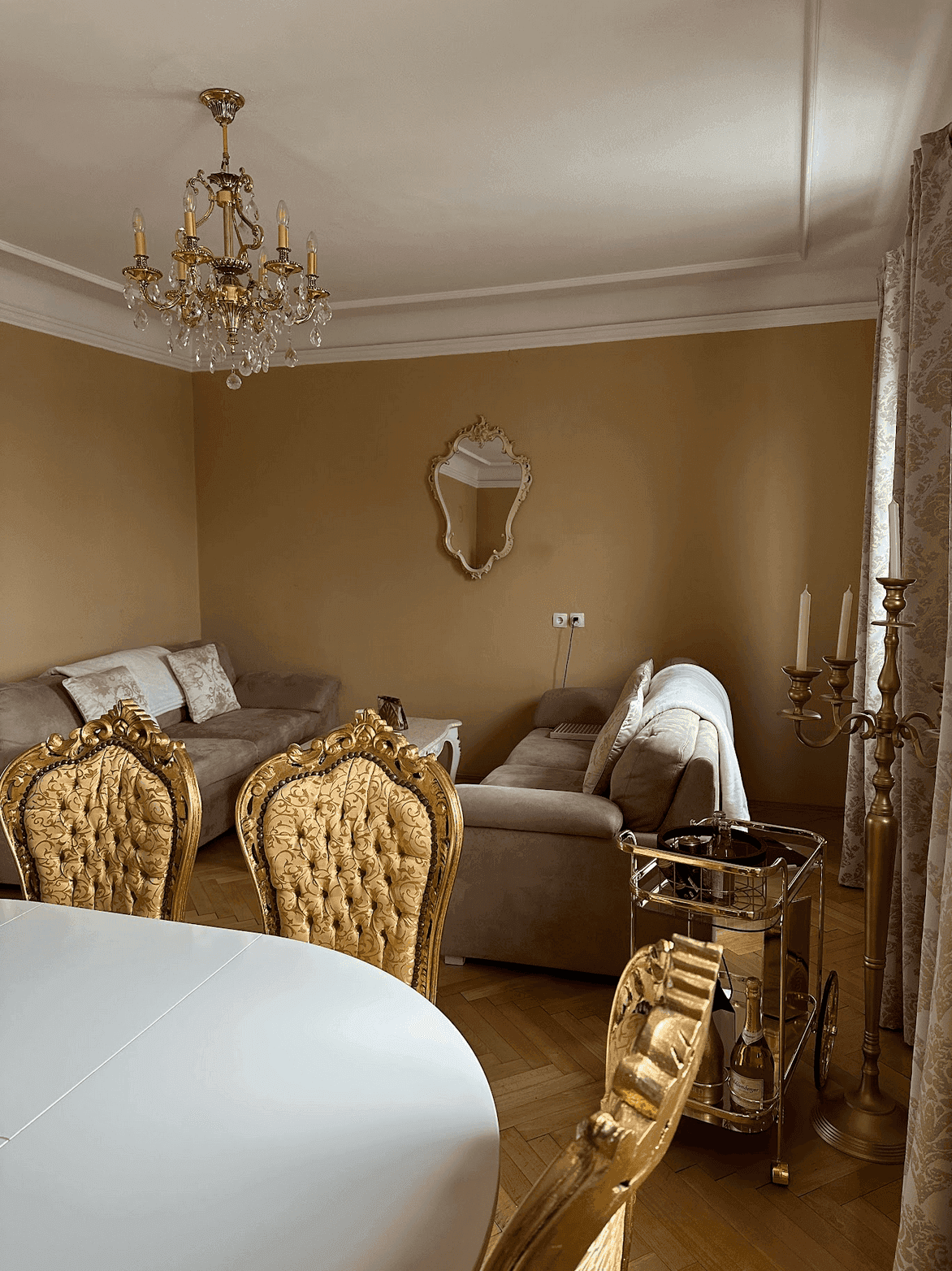 Heller Wohn-Essbereich mit beigen Sofas, goldenen Stühlen und einem eleganten Kronleuchter.