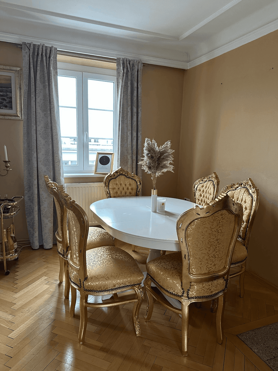 Eleganter Essbereich mit ovalem Tisch, goldenen Stühlen und Fischgrätparkettboden unter einem Fenster.