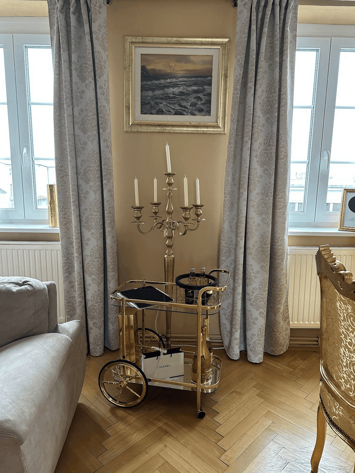 Heller Wohnbereich mit großen Fenstern, Kerzenleuchter und einem goldenen Servierwagen.