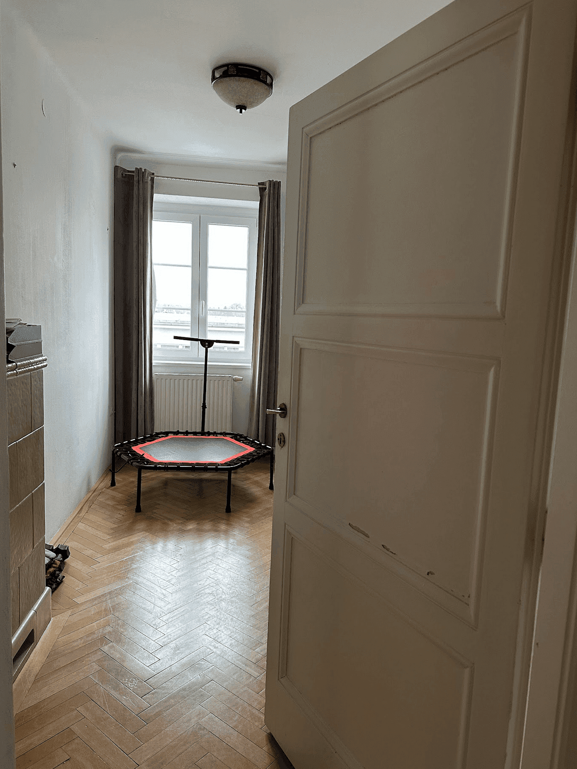 Kleines Zimmer mit Fischgrätparkett, Fenster und einem Trampolin, ideal als Kinder- oder Spielzimmer.