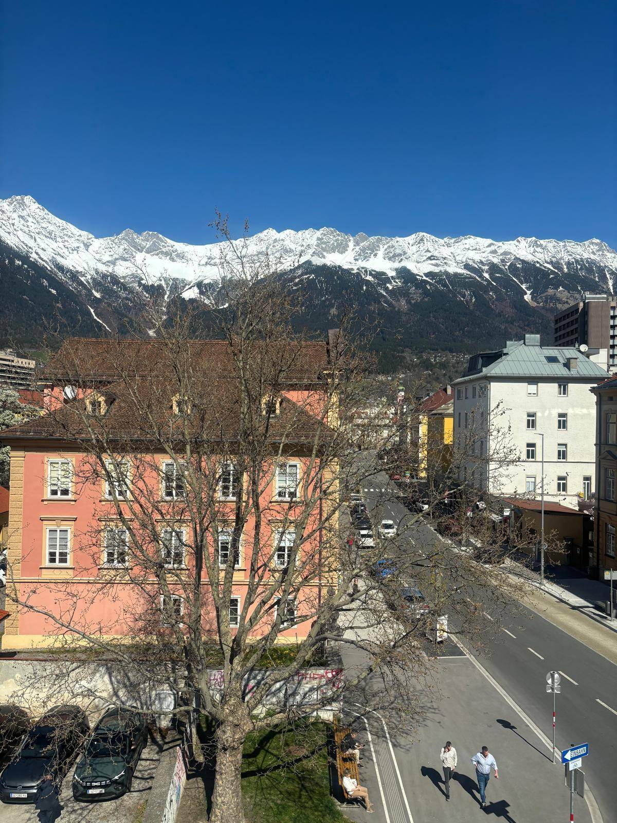 Panoramablick auf schneebedeckte Berge, eine Stadtstraße und umliegende Gebäude unter blauem Himmel.