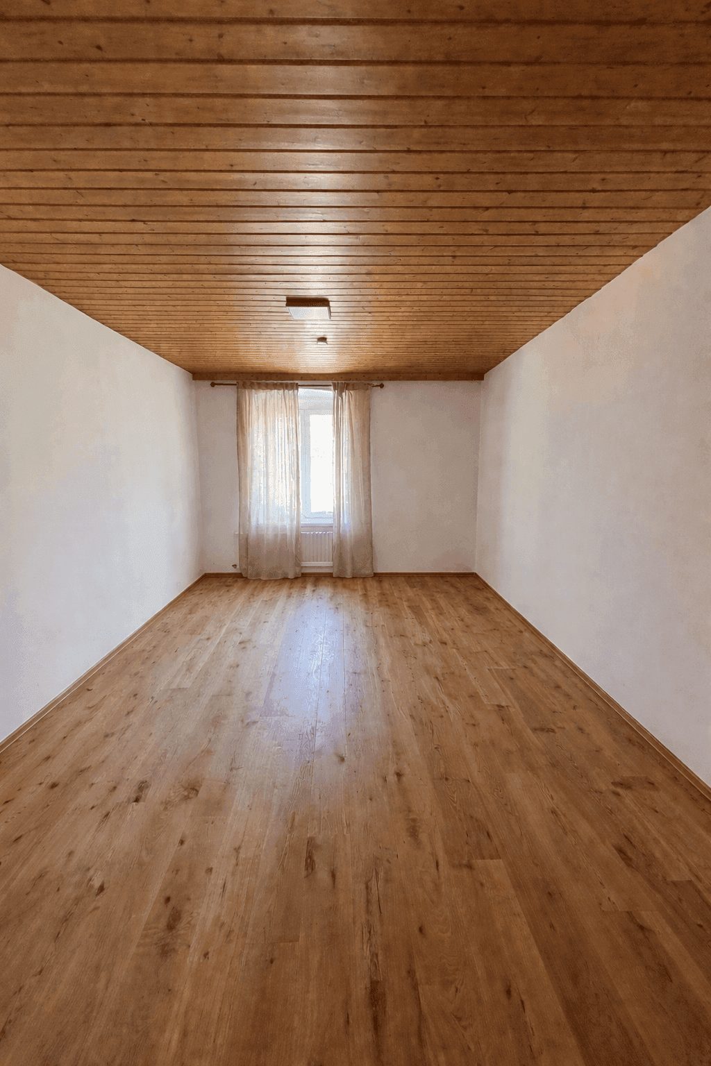 Leerer Raum mit Holzdeckenverkleidung, Parkettboden und einem Fenster mit Vorhängen.